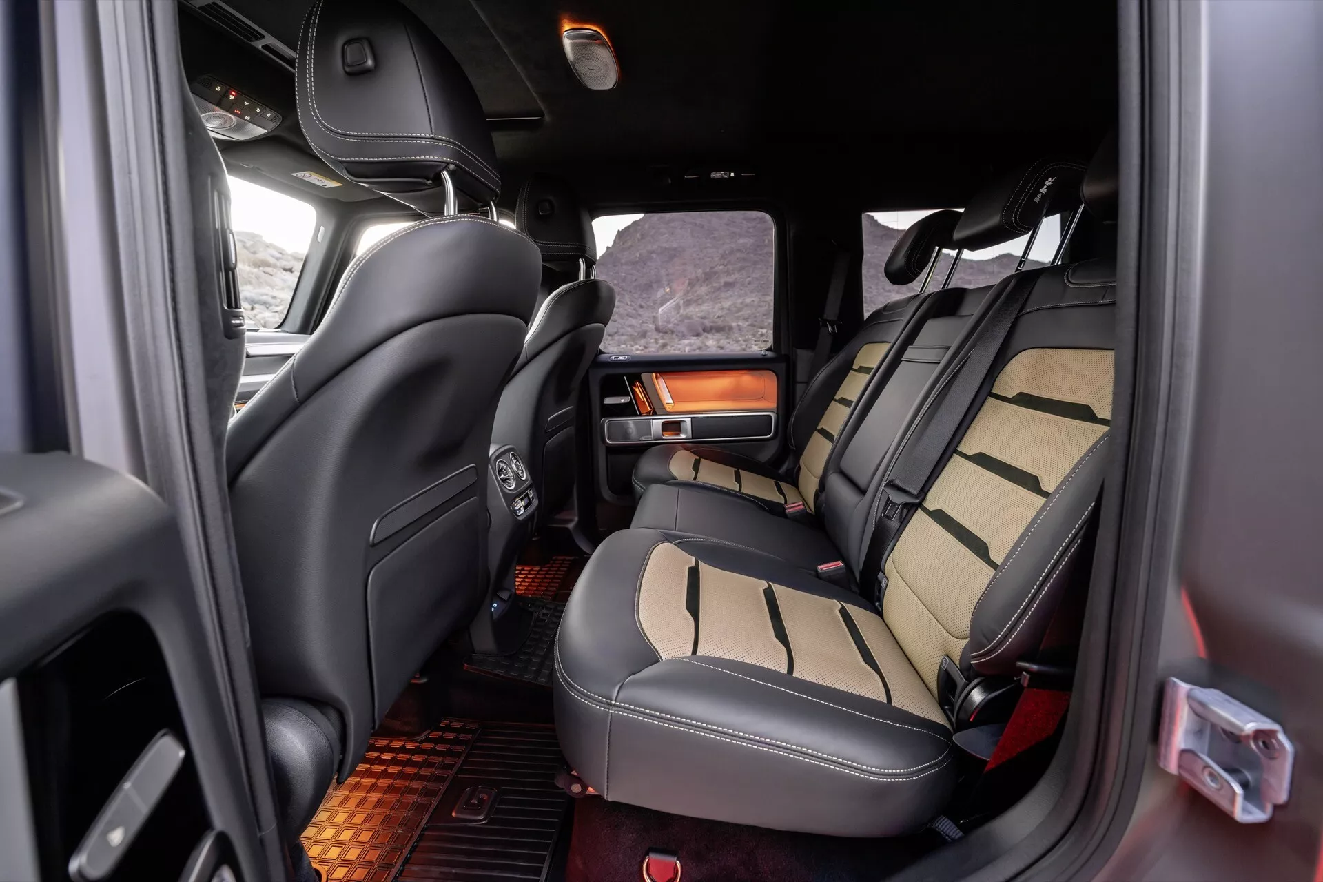 Vue intérieure de la Mercedes-Benz Classe G (G-Klasse) 2024 montrant la zone des sièges de la deuxième rangée avec sièges revêtus de cuir bicolore, accoudoir repliable avec porte-gobelet, sièges de la troisième rangée visibles et détails luxueux incluant des éléments en bois et un éclairage d'ambiance