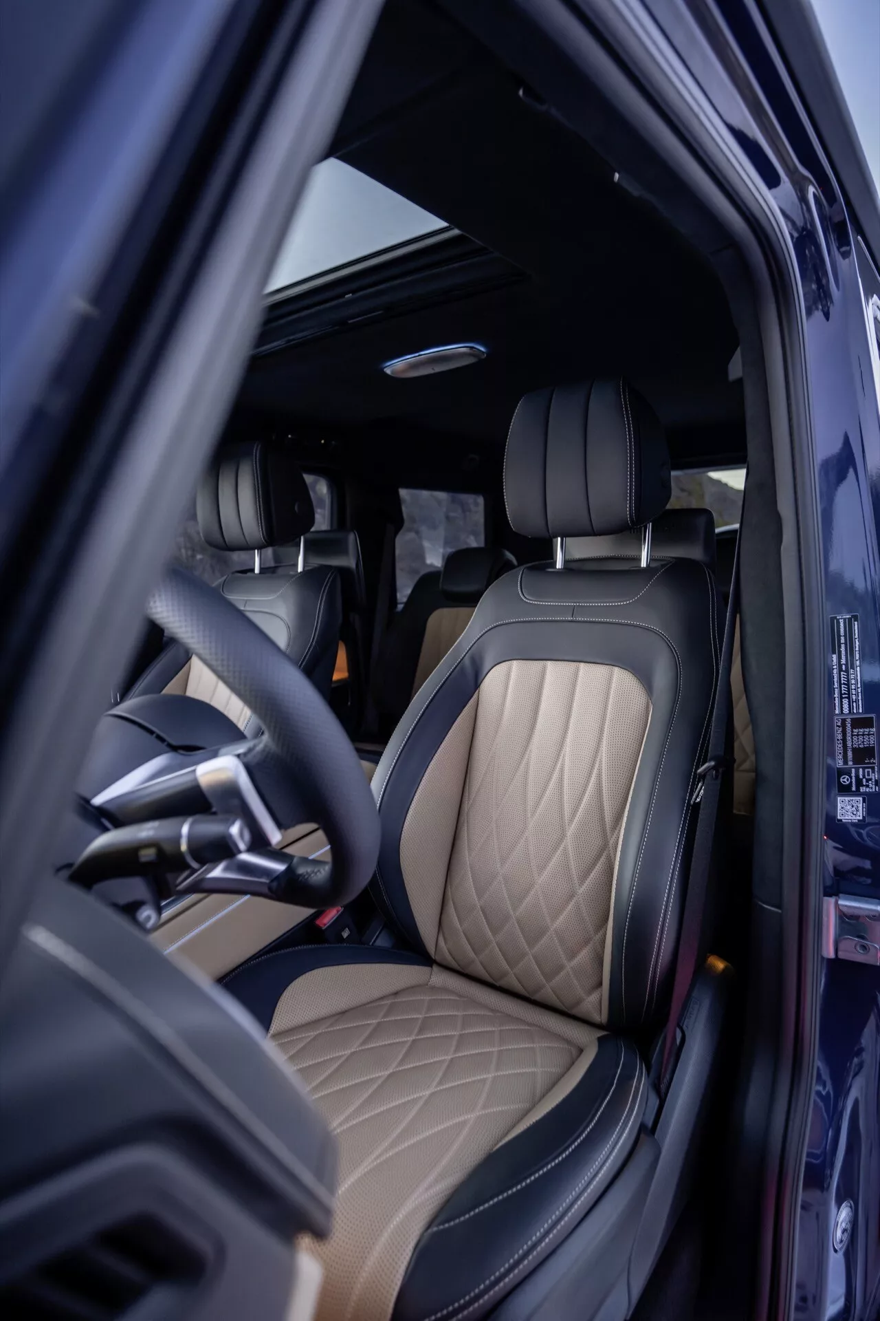Vue de l'intérieur de la Mercedes-Benz Classe G 465 2024 côté conducteur avec porte ouverte, mettant en avant une sellerie de luxe et des matériaux haut de gamme, volant ergonomique et éléments de design sophistiqués