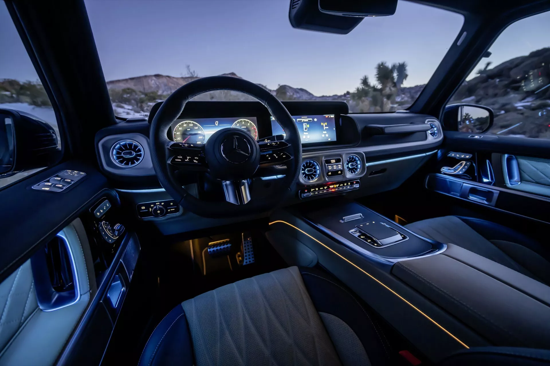 Vue intérieure de la Mercedes-Benz G-Klasse 2024, mettant en avant la conception du cockpit depuis la perspective du passager arrière, présentant le volant sportif multifonction, le tableau de bord avec cluster d'instruments numériques, système d'infodivertissement à écran large, contrôle du climat, éclairage intérieur ambiant, sièges avant luxueux, sélecteur de vitesse, porte-gobelets, conception de porte avec commandes électriques et matériaux de finition premium.
