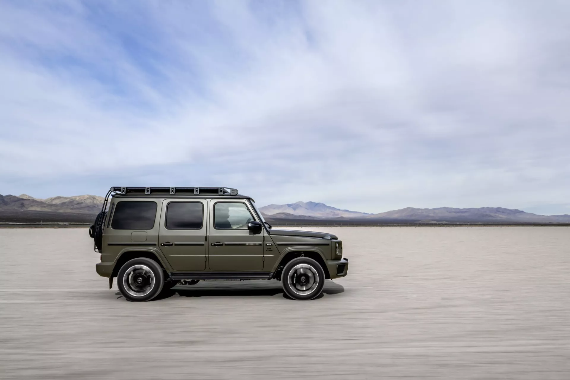 Mercedes-Benz G-Klasse 2024 en style SUV militaire se déplaçant sur des étendues salines, montrant le profil latéral et frontal avec un flou de mouvement, mettant en avant son design robuste, sa capacité tout-terrain et ses fonctionnalités traditionnelles sans technologie moderne visible