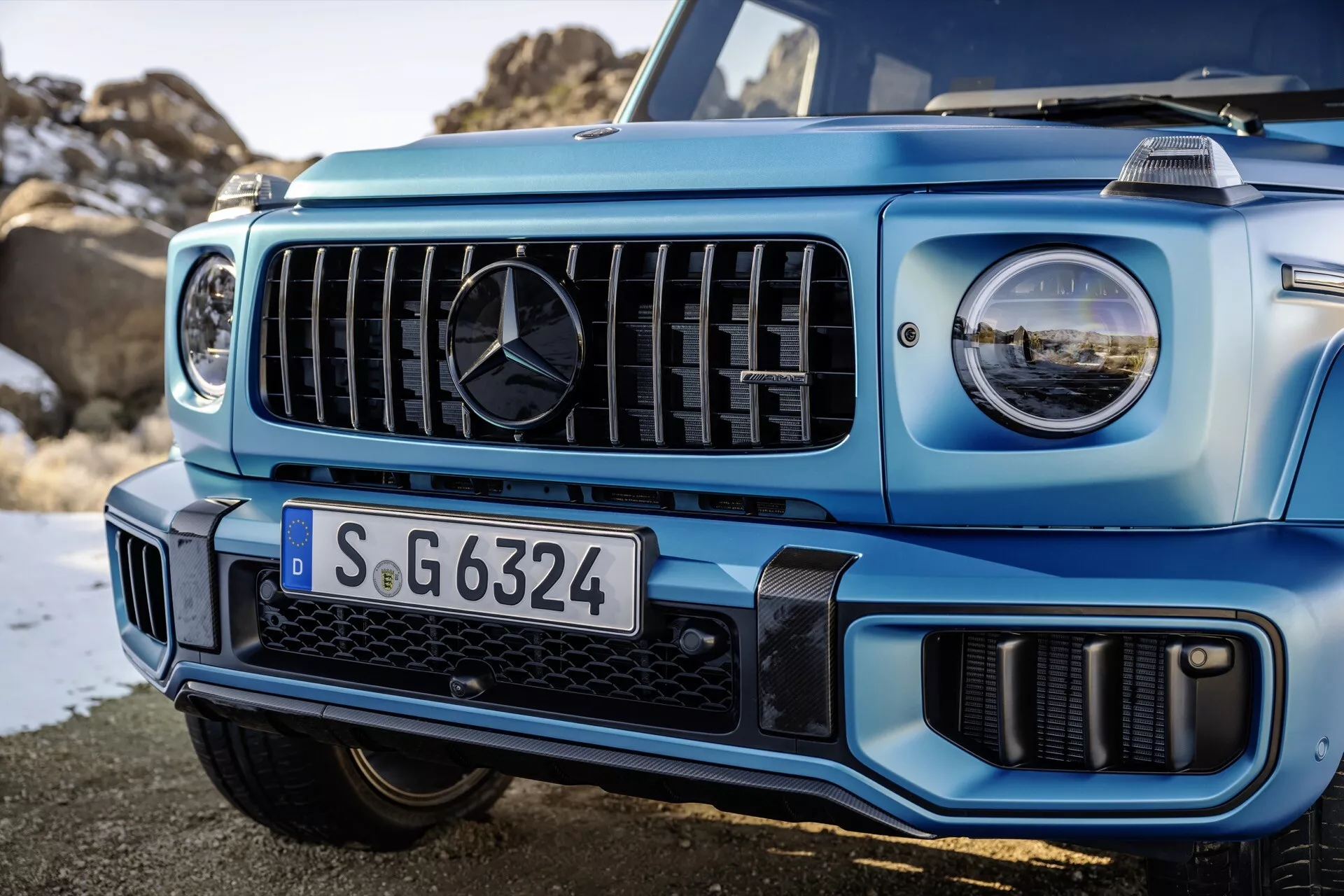 Vue rapprochée du côté avant gauche d'un Mercedes-Benz Classe G de 2024 en bleu clair, mettant en évidence la grille avec des lamelles verticales et des phares LED, illustrant la technologie d'éclairage avancée et la capacité tout-terrain du SUV
