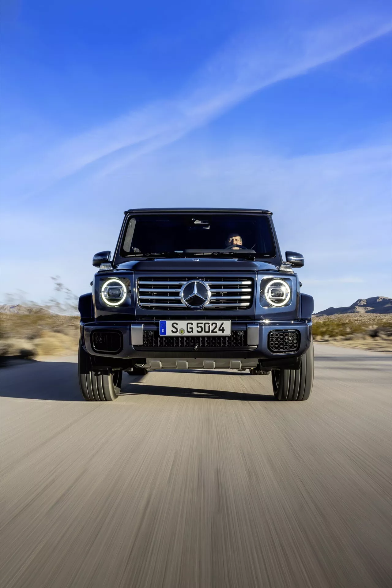 SUV Mercedes-Benz Classe G 2024 en mouvement, capturé d'un angle frontal bas soulignant son design agressif et ses fonctionnalités avancées telles que la grille emblématique, phares ronds, grandes prises d'air et jantes en alliage contre un fond désertique
