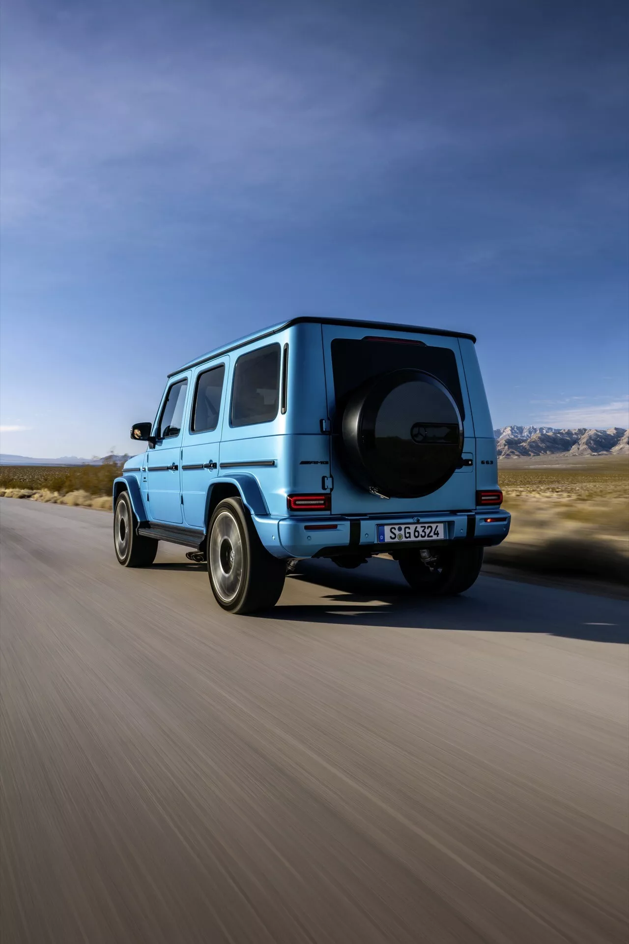 SUV Mercedes-Benz Classe G 2024 en bleu vif, capturé sous un angle arrière trois quarts montrant le profil droit, la roue arrière, la porte du passager arrière, une partie du toit et la roue de secours montée, sur un fond de paysage avec ciel clair et montagnes, suggérant aventure et performances