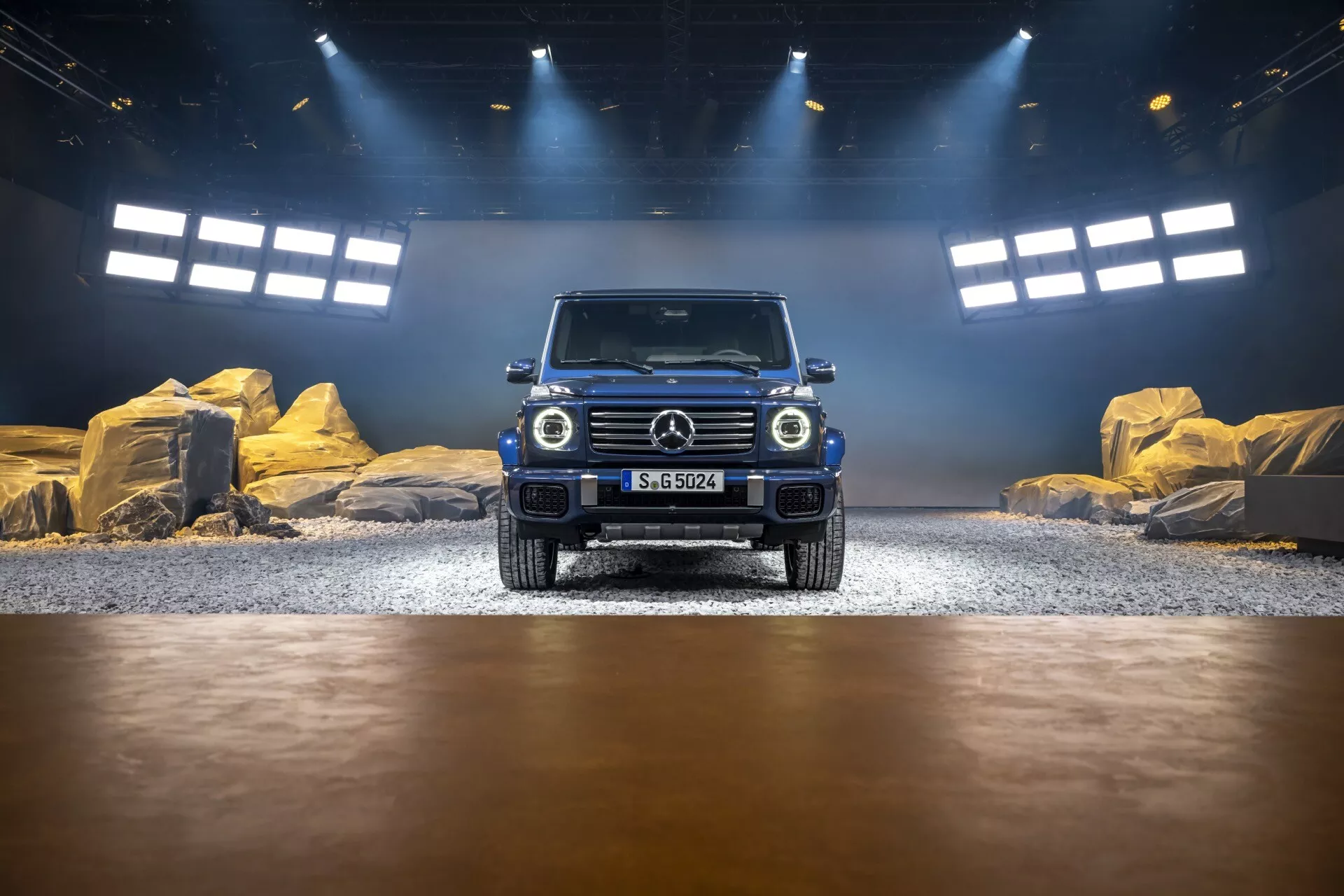 Mercedes-Benz Classe G 2024 présentée dans un cadre intérieur simulant un environnement tout-terrain, vue de l'angle avant mettant en évidence la grille, les phares LED et la conception du pare-chocs avant, avec des effets de lumière de studio et naturelle soulignant sa peinture métallique et ses capacités tout-terrain