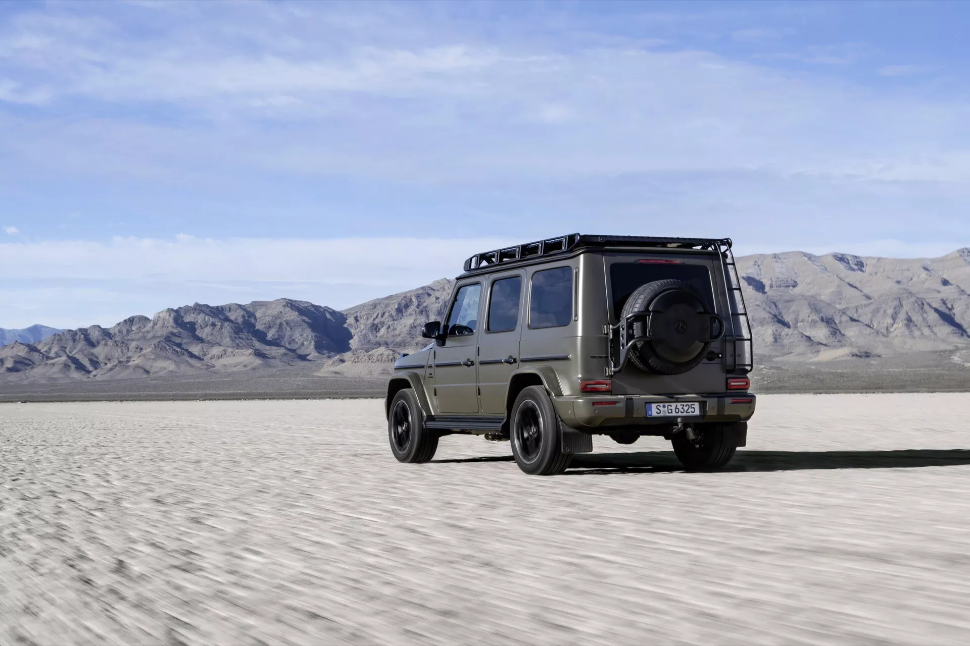 SUV Mercedes-Benz G-Klasse 2024 capture son profil latéral avec une touche de l'arrière, montrant des capacités tout-terrain sur un terrain désertique, avec des barres de toit, des pneus robustes et une roue de secours montée sur la porte arrière