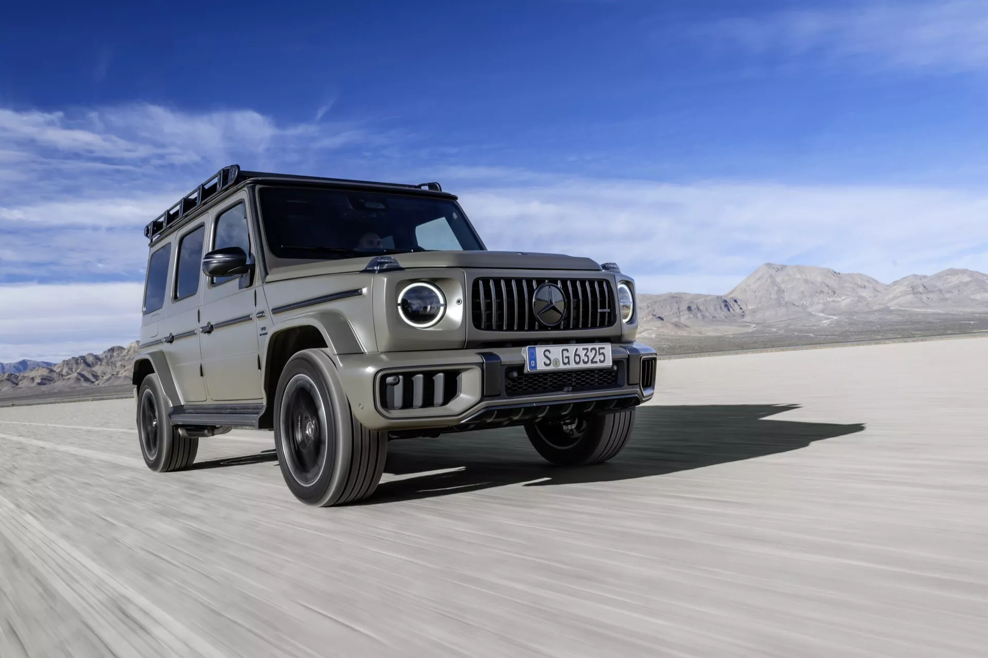 SUV Mercedes-Benz Classe G 2024 en argent, démontrant ses capacités tout-terrain sur un terrain désertique, avec une prise de vue dynamique et sous un angle bas soulignant son design robuste, ses phares ronds, sa grille avant et son sillage de poussière.