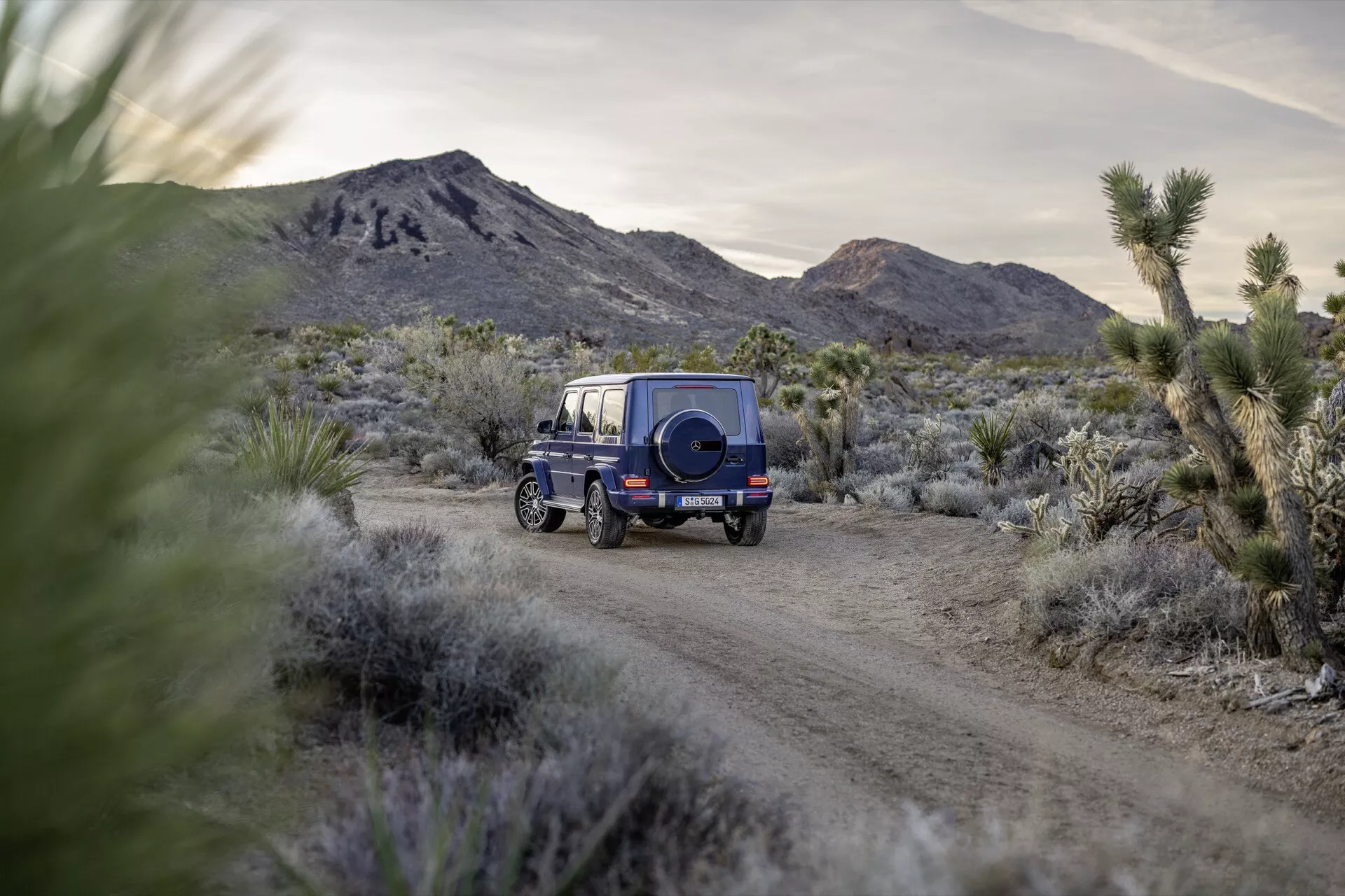 SUV bleu Mercedes-Benz G-Klasse (Classe G) 2024 dans une vue arrière aux trois quarts roulant sur une route de terre avec une roue de secours sur le hayon et un paysage désertique à l'aube ou au crépuscule