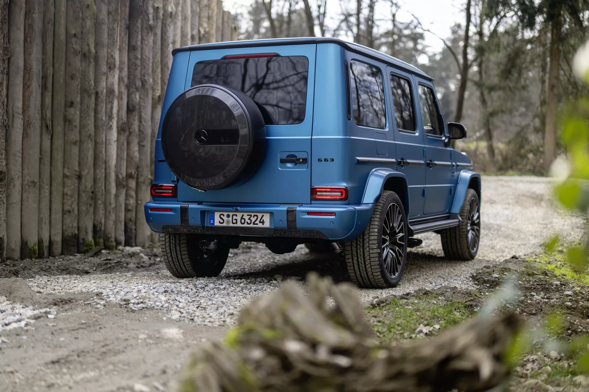 Vue arrière du SUV bleu Mercedes-Benz Classe G (G 465) 2024 soulignant son design robuste et carré, roue de secours montée, feux arrière LED, grande garde au sol et roues en alliage sur un chemin de gravier entouré par la nature