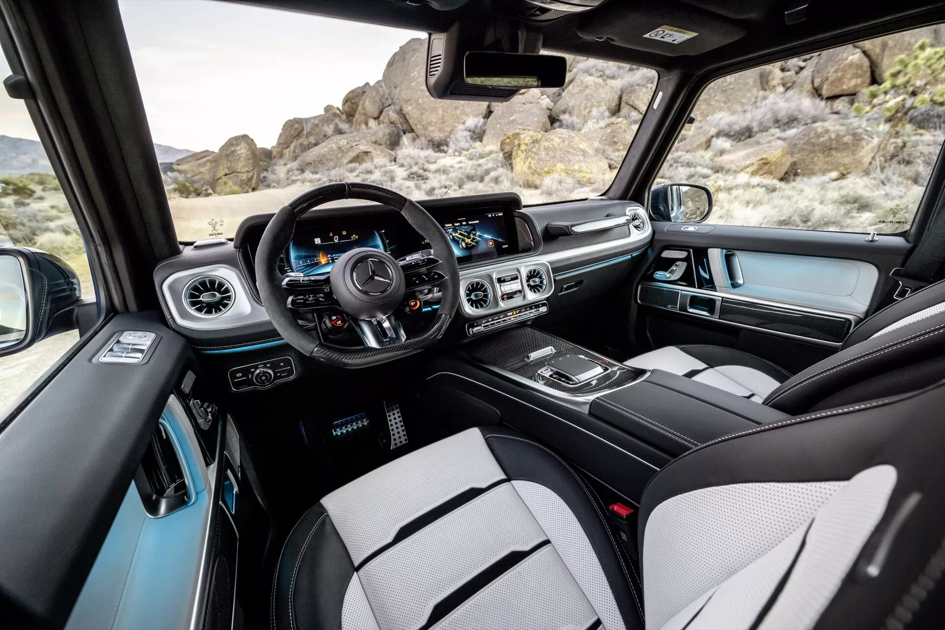 Intérieur du Mercedes-Benz G-Klasse 2024 vu depuis la zone des sièges arrière, montrant le tableau de bord central et côté conducteur, commandes sur la porte du conducteur, volant avec boutons multifonctions, panneau d'instruments, système d'infodivertissement, contrôles du climat, console centrale et vue partielle sur le toit ouvrant.