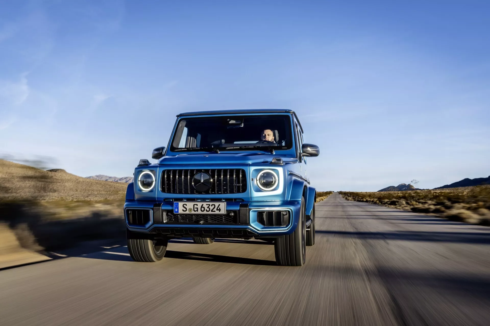 SUV de luxe Mercedes-Benz Classe G 2024 en mouvement, capturé depuis une perspective frontale avec technique de prise de vue de tracking montrant la grille détaillée, les phares LED et les fonctionnalités ADAS, sur une route désertique sous un ciel bleu clair