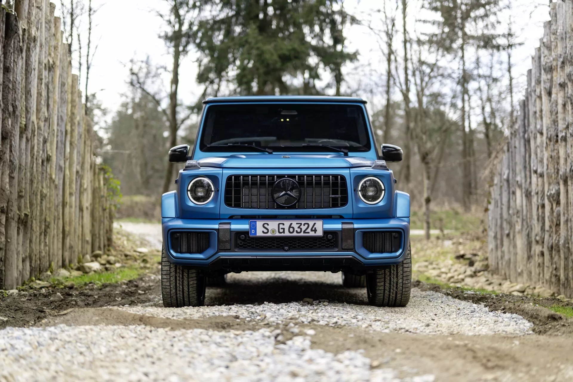 Vue de face d'un SUV bleu Mercedes-Benz G-Klasse 2024 sur un chemin de gravier, entre des rangées de poteaux en bois, soulignant le design moderne du véhicule et les phares LED.