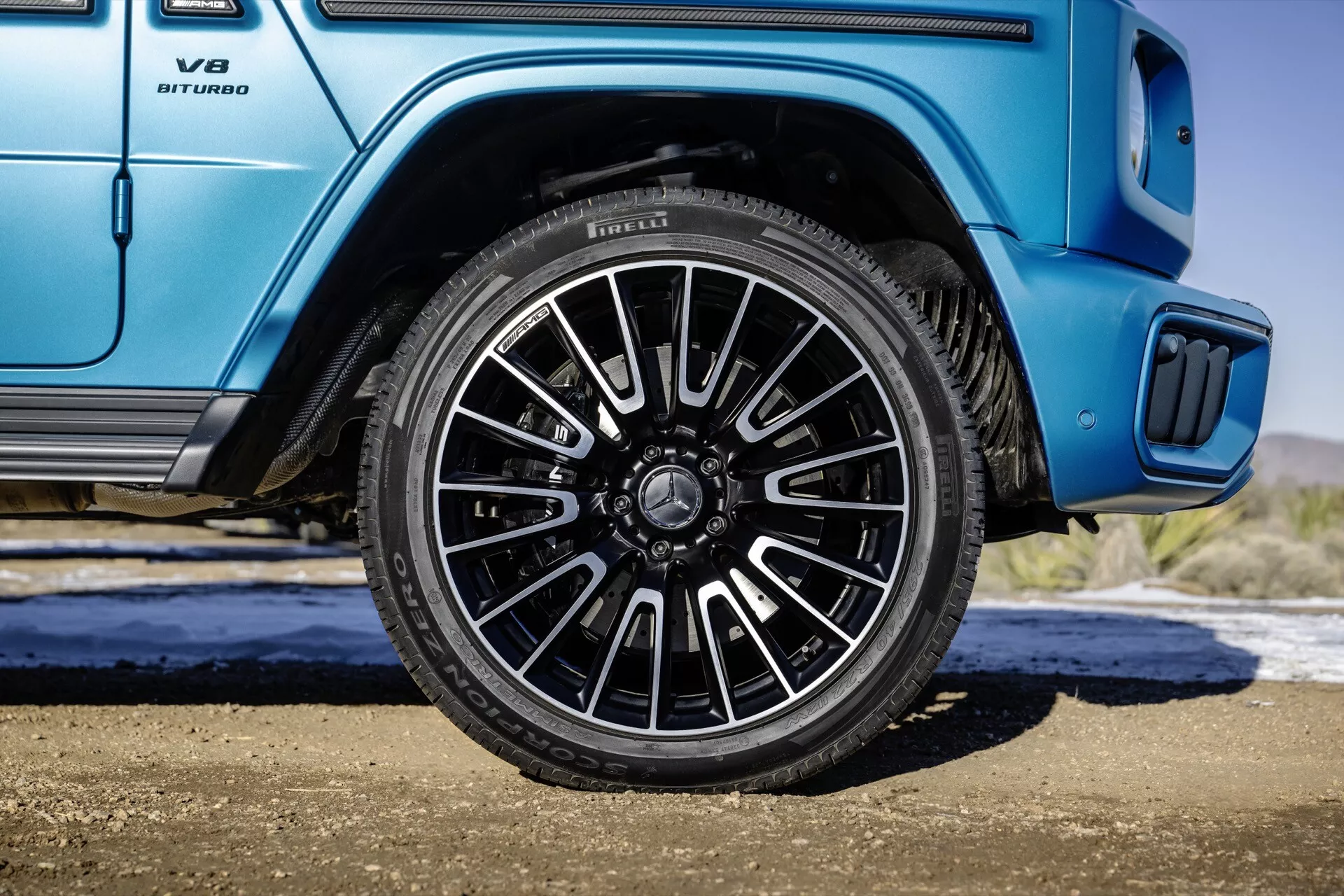 Mercedes-Benz Classe G (G 465) 2024 présentant la roue arrière côté passager avec un design à multiples branches et une finition sombre contre une carrosserie bleue vibrante, incluant un gros plan sur l'emblème V8 BITURBO et le détail du marchepied