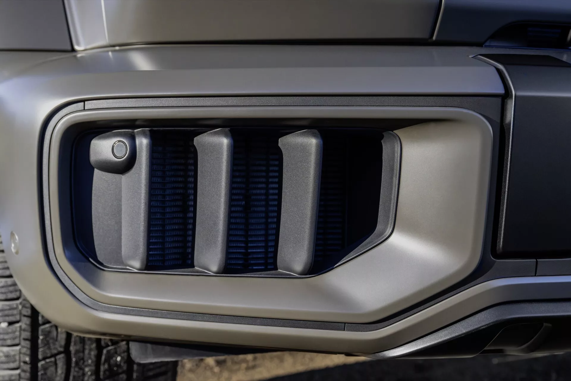 Gros plan sur le pare-chocs avant de la Mercedes-Benz G-Klasse 2024 présentant une grille de ventilation avec capteur intégré, soulignant la technologie avancée et l'esthétique design moderne.