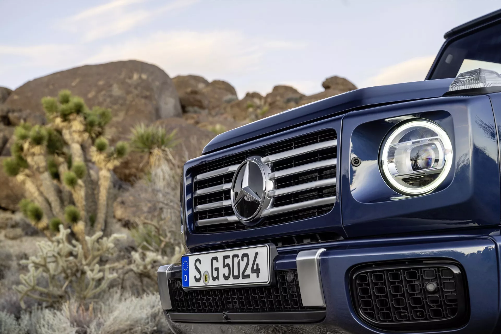 Vue rapprochée du côté avant gauche du Mercedes-Benz G-Klasse 2024 mettant en lumière la grille distinctive avec emblème central, une partie du phare LED avancé, sur un arrière-plan naturel extérieur suggérant des capacités tout-terrain robustes