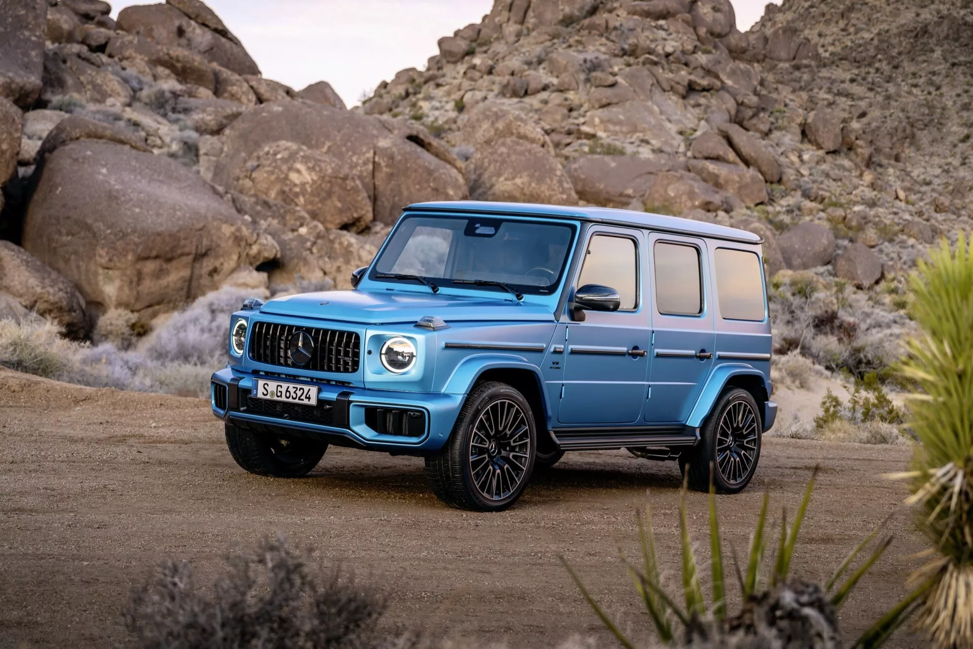 SUV de luxe Mercedes-Benz G-Klasse 2024 en bleu clair, présentant sa grille distinctive, ses grandes prises d'air, et ses phares LED sous un angle avant de trois quarts sur un sol en terre battue avec un arrière-plan de désert rocheux, mettant en avant ses capacités tout-terrain et ses caractéristiques de design moderne.
