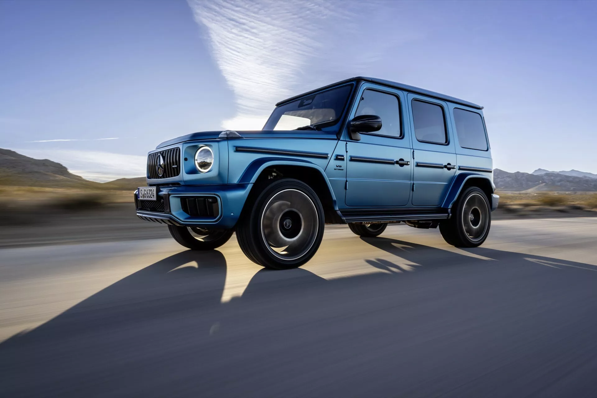 SUV de luxe Mercedes-Benz G-Class 2024 capturé en mouvement depuis une vue latérale dynamique et en contre-plongée, mettant en valeur son design robuste et carré, peinture bleue avec garnitures noires, phares ronds, feux de jour à LED et jantes en alliage noires contre un arrière-plan flou à grande vitesse.