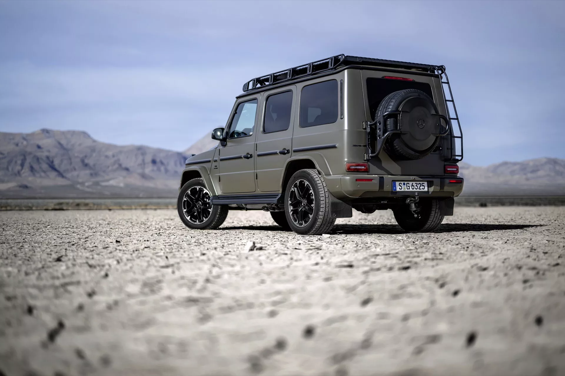 SUV Mercedes-Benz Classe G 2024 garé sur un lit de lac désertique, montrant son design robuste avec porte-bagages, roue de secours montée à l'arrière et jantes en alliage stylisées sous un ciel clair avec des montagnes lointaines.