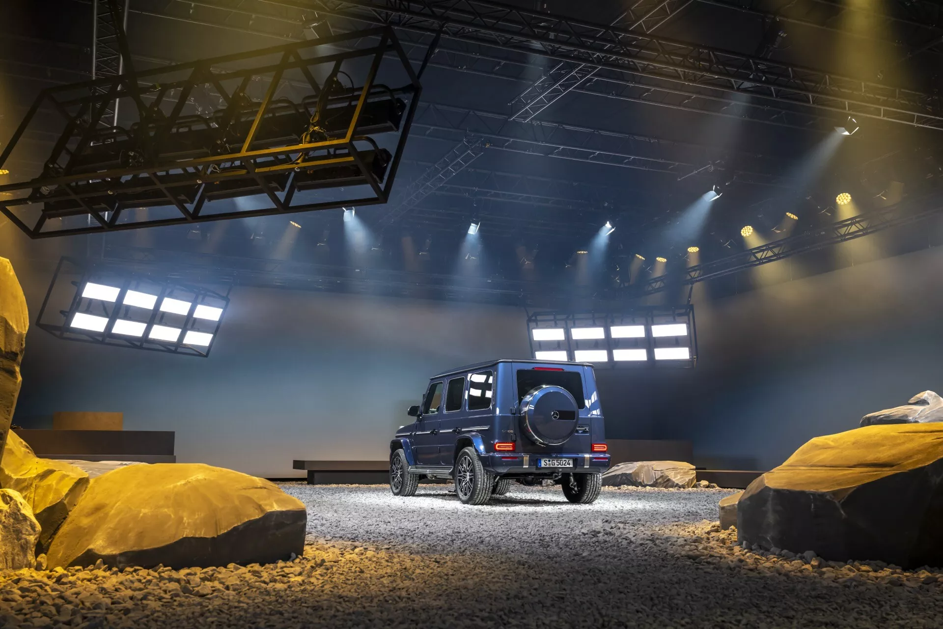 SUV noir Mercedes-Benz Classe G (G-Klasse 465) 2024 vu de trois quarts arrière montrant les capacités tout-terrain avec roue de secours, feux arrière LED et design de luxe dans un environnement intérieur simulé tout-terrain avec gravier et structures semblables à des roches sous un éclairage artificiel dramatique