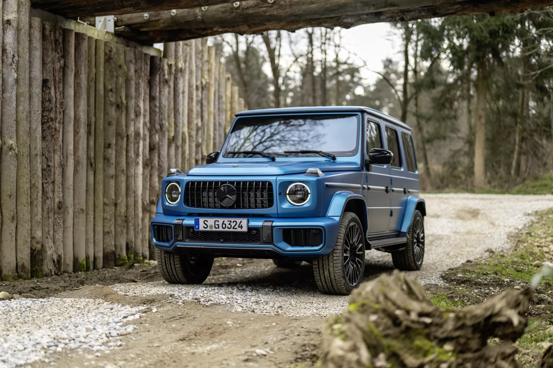 Vue de trois quarts avant d'un SUV Mercedes-Benz Classe G 2024 en bleu, mettant en évidence la grille avant, les phares LED, le pare-chocs avant et les roues en alliage, sur un fond rustique hors route avec éclairage doux.