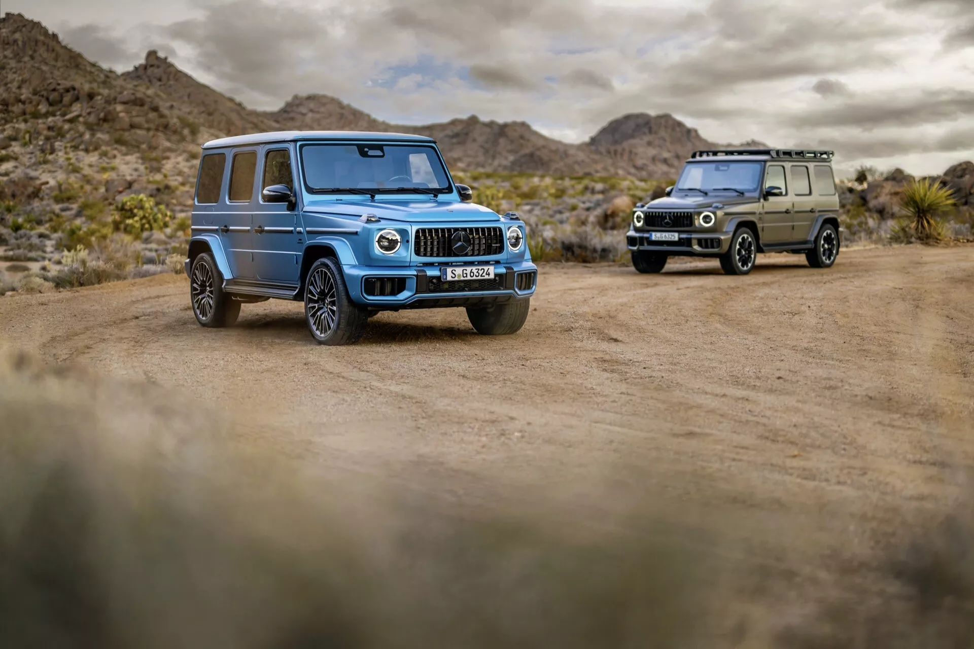 Mercedes-Benz Classe G 465 2024 en bleu clair, exposée de manière proéminente dans un paysage désertique sur une route en terre, montrant des phares LED, des lamelles de grille chromées verticales, de grandes roues alliage à multiples branches avec des pneus tout-terrain et des fonctionnalités technologiques avancées, avec une seconde Classe G en vert olive visible en arrière-plan.