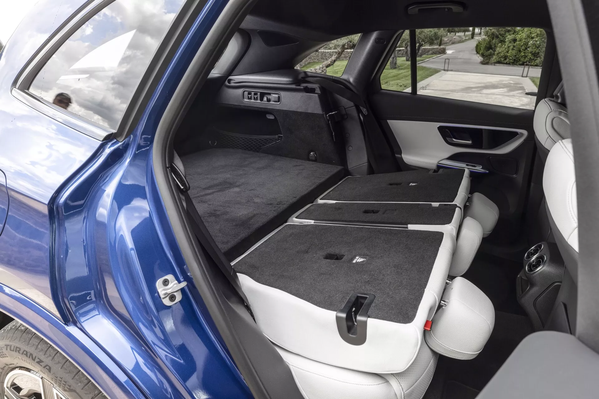 Vue partielle du Mercedes GLC 254 2022 mettant en avant l'espace de chargement intérieur modulable avec les sièges arrière rabattus, vue en diagonale depuis l'arrière droit. L'extérieur bleu foncé avec la porte passager arrière ouverte, l'arche de roue arrière visible et des caractéristiques technologiques telles que le contrôle des fenêtres sur le panneau de porte et la grille du haut-parleur sont partiellement visibles, renforçant l'utilité et le design de la voiture.