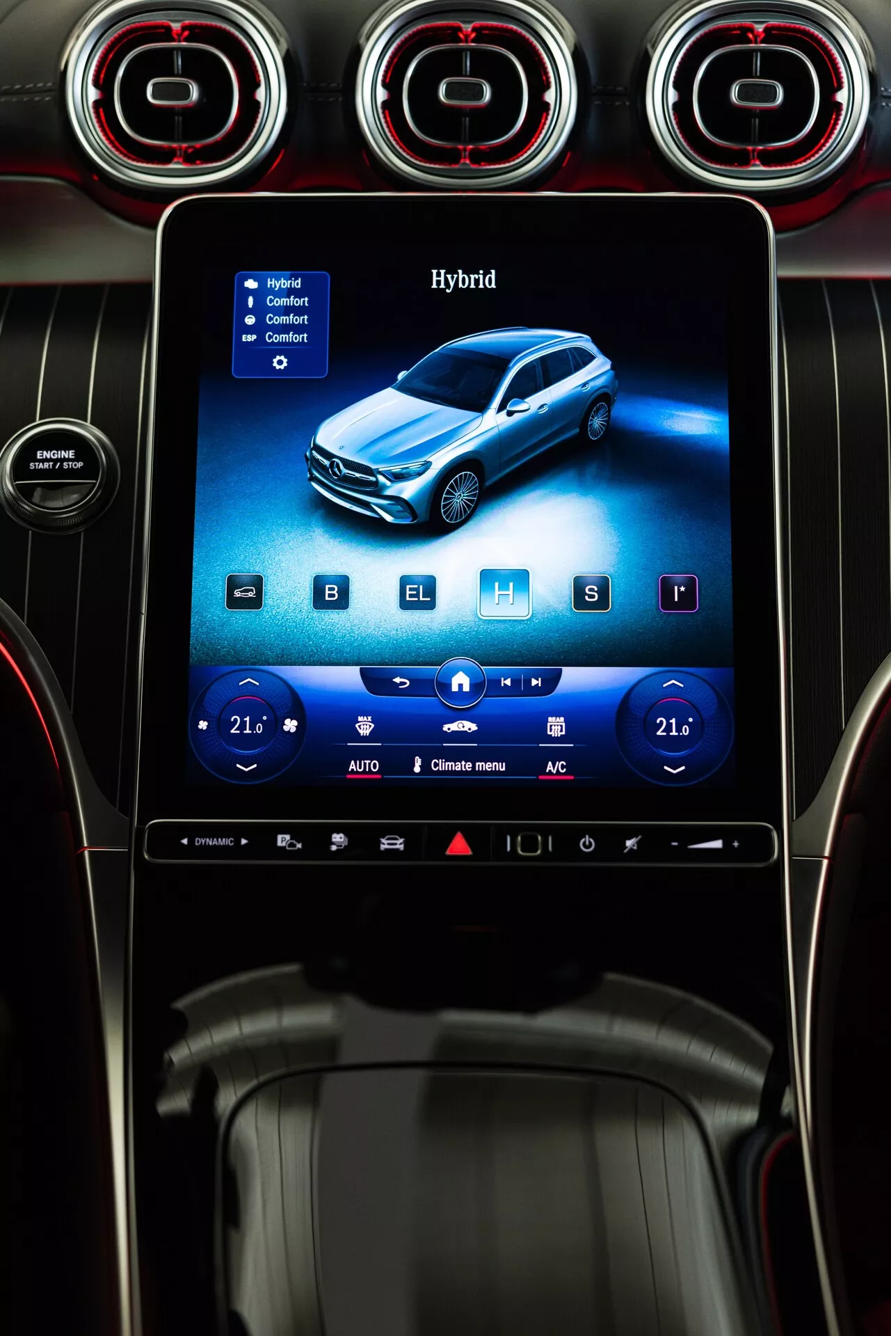Vue rapprochée de la console centrale du Mercedes GLC 254 de 2022, avec un système d'infodivertissement à écran tactile vertical encadré par trois aérateurs circulaires avec des anneaux rouges accentués. L'écran affiche un véhicule hybride bleu, avec des commandes de climatisation numériques réglées à 21 degrés Celsius de chaque côté, indiquant une climatisation à double zone. Des icônes pour les sièges chauffants, le refroidissement et les options de dégivrage sont visibles, ainsi qu'une barre d'accès rapide en bas. La console est conçue avec des accents en métal brossé, des surfaces noires brillantes et des boutons minimalistes, soulignant un intérieur moderne et luxueux.