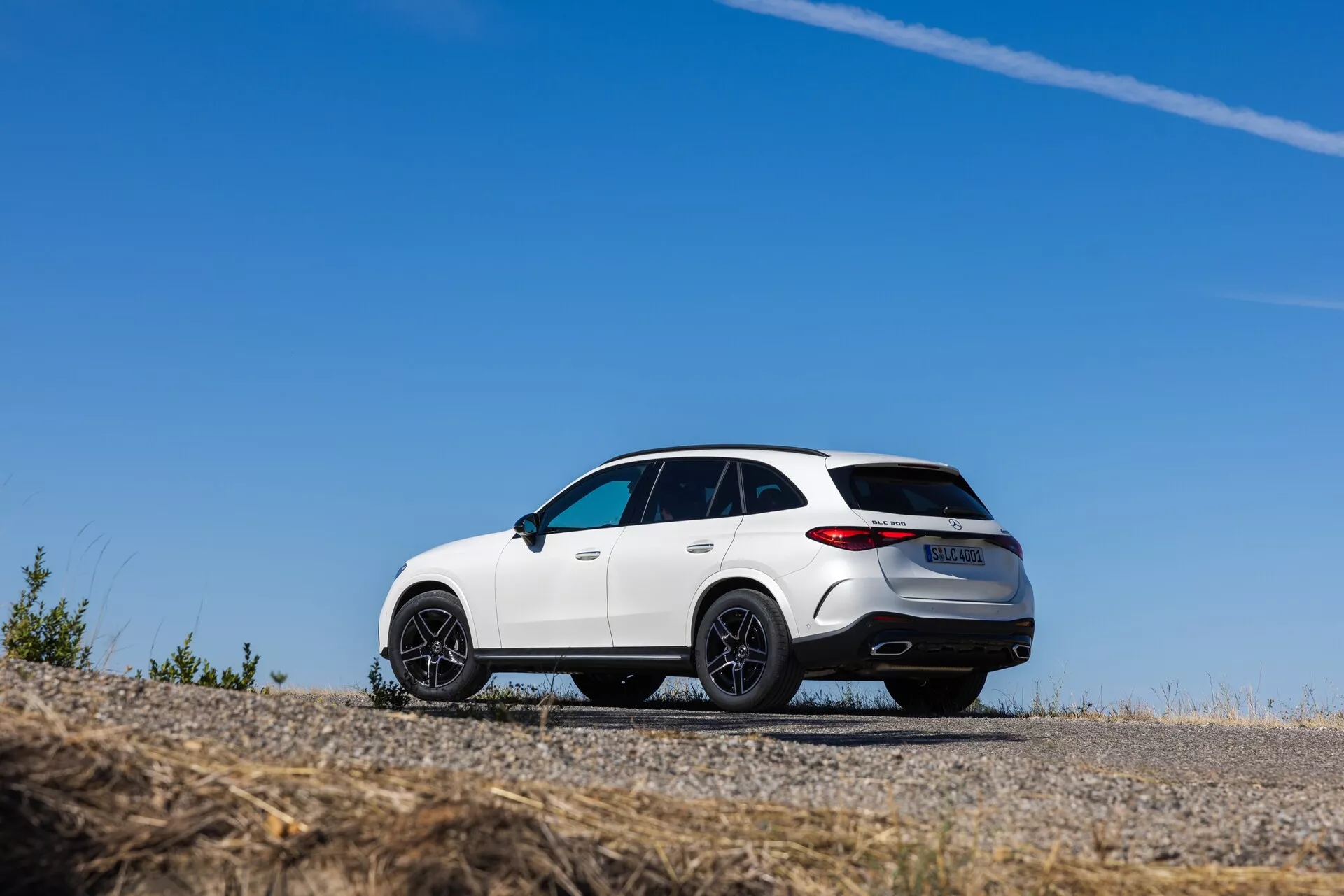 Vue arrière trois-quarts en contre-plongée d'un SUV Mercedes GLC 2022 blanc, garé sur une surface en asphalte, mettant en avant son design sportif avec une ligne de toit inclinée, de grandes jantes alliage noires multi-rayons et des feux arrière à LED. Le véhicule présente des accents chromés, des sorties d'échappement doubles, un aileron arrière, un toit panoramique et un diffuseur arrière, soulignant sa construction de qualité supérieure et son allure sportive.