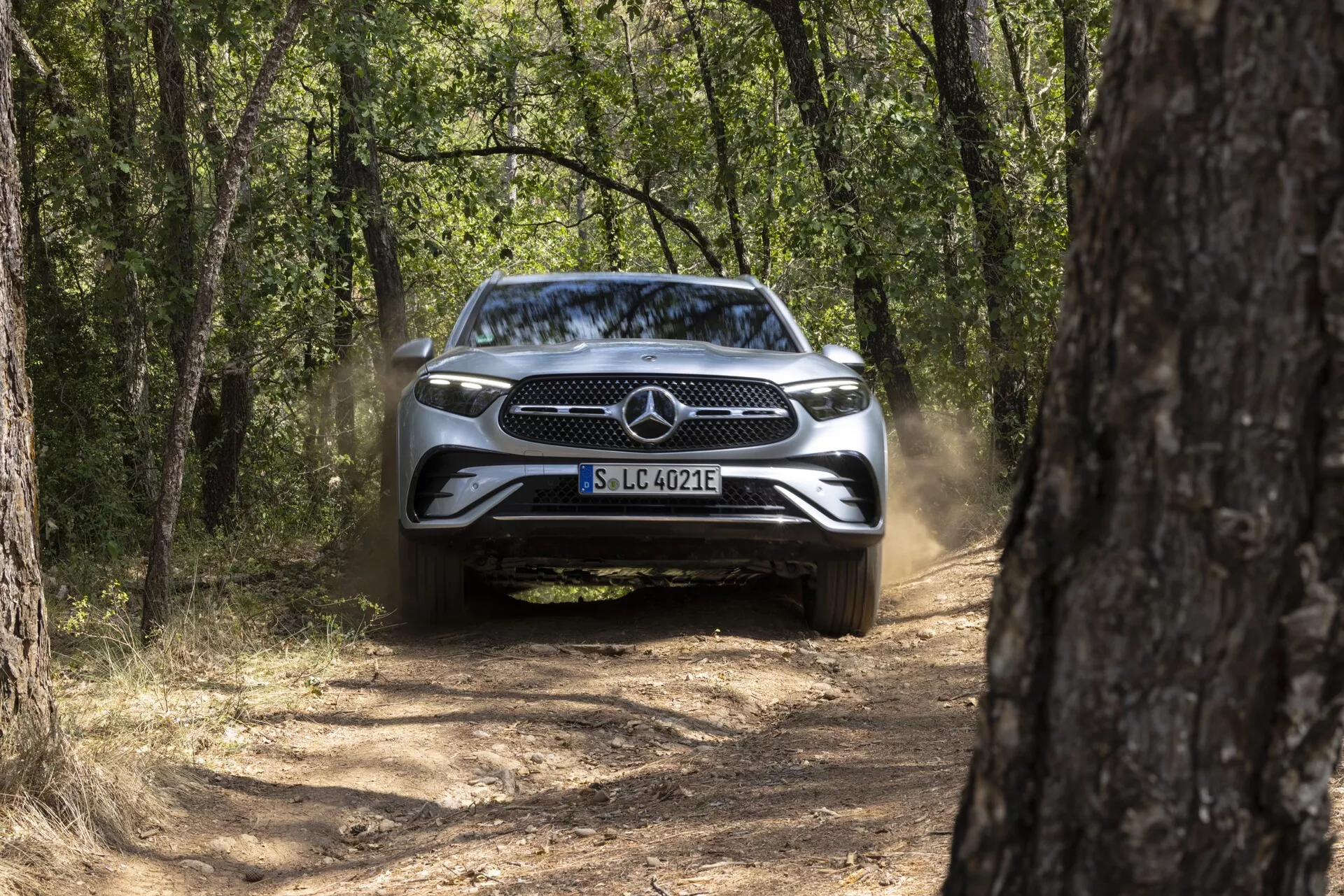 Vue avant d'une Mercedes GLC 2022 (254) sur un chemin de terre étroit, encadrée par des arbres, phares éteints, poussière derrière, journée ensoleillée avec lumière naturelle.