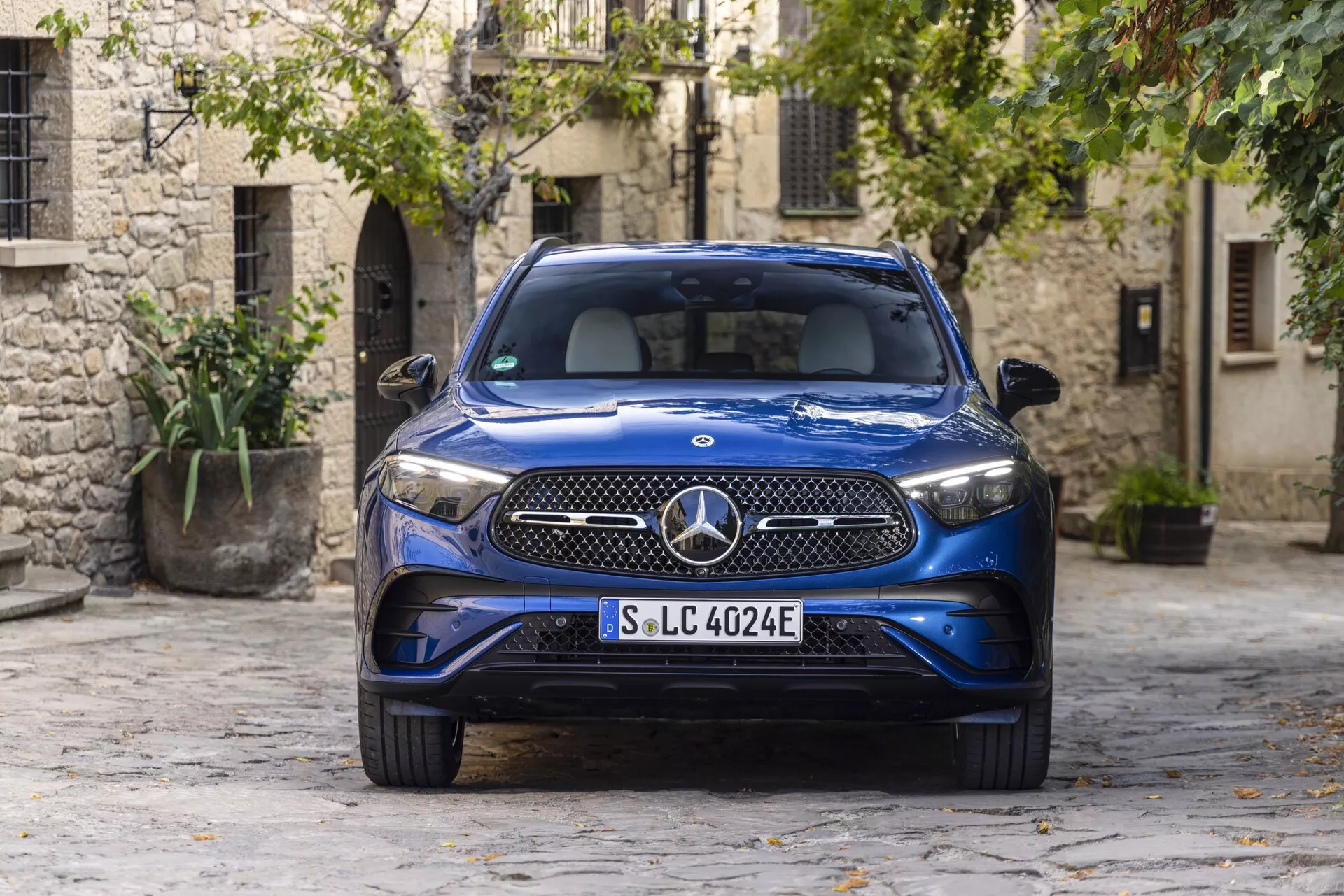 SUV de luxe Mercedes GLC 254 bleu 2022 garé dans une cour pavée pittoresque d'une vieille ville, présentant des phares LED, une grille distinctive avec un grand emblème Mercedes-Benz, un badge EQ indiquant une nature électrique ou hybride rechargeable, une carrosserie métallique élégante et des bouches d'aération intégrées dans le pare-chocs avant. Capturé sous un angle frontal, mettant en valeur le design automobile moderne contre des bâtiments en pierre traditionnels et de la verdure.