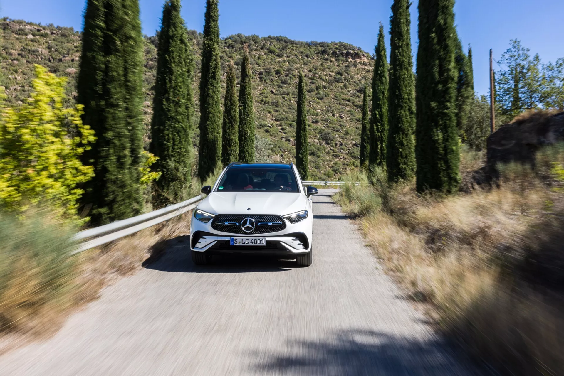 Image dynamique en perspective frontale d'une Mercedes GLC 2022 (254) roulant vers la caméra sur une route étroite et ensoleillée bordée de grands arbres, montrant la face avant complète, y compris la calandre avec l'emblème Mercedes-Benz, les phares LED élégants, le capot, le pare-brise et une partie du toit. Le léger flou des roues et le fond strié créé par la technique du flou de mouvement met en évidence le mouvement de la voiture et les fonctionnalités avancées telles que la technologie des phares LED éventuelle, la conception actualisée de la calandre avec des éléments aérodynamiques actifs ou des capteurs pour les systèmes d'assistance à la conduite, et probablement des capteurs de stationnement ou des caméras.