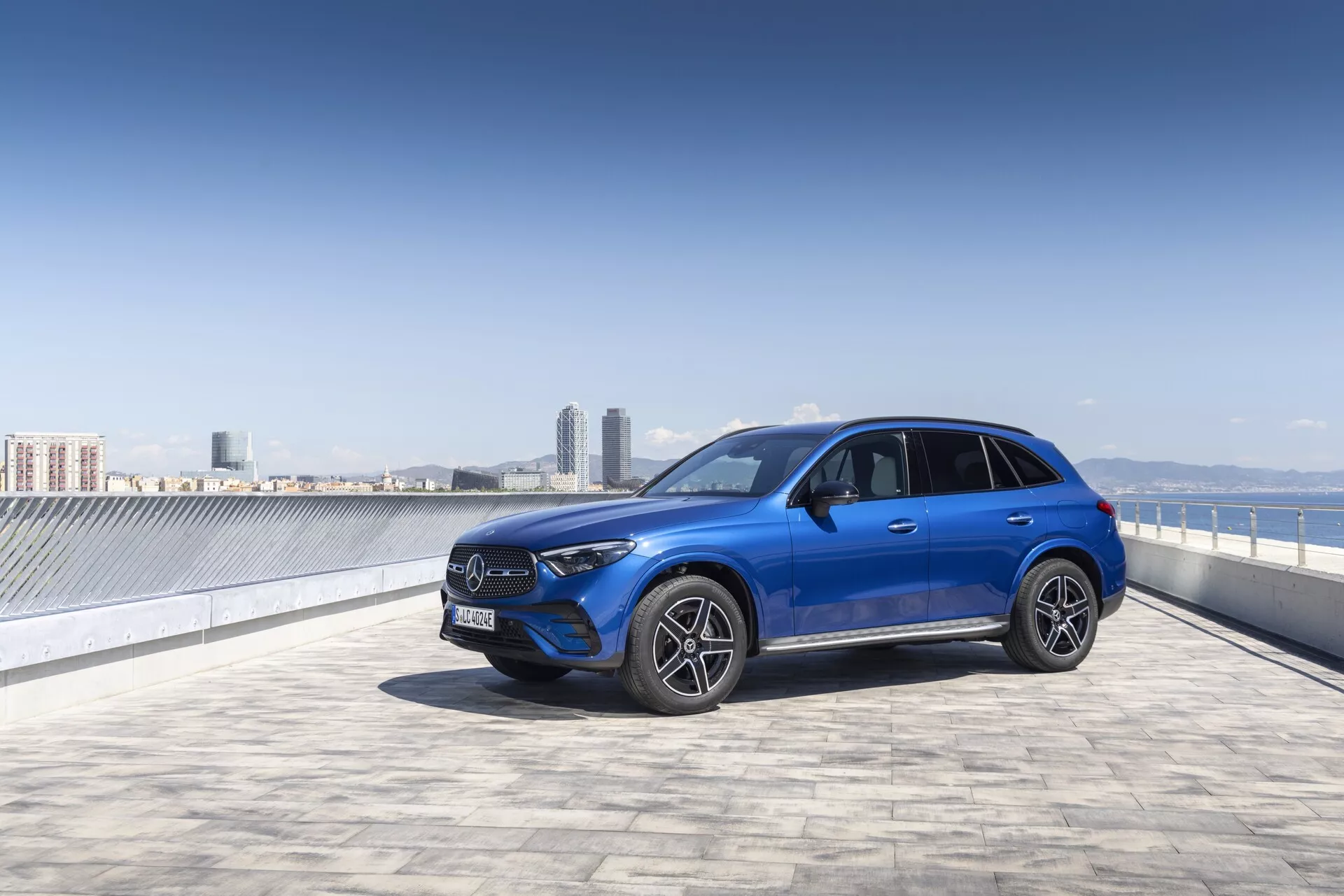 Mercedes GLC 2022 SUV bleu vif sur une aire de stationnement en toiture avec un ciel dégagé et une skyline urbaine en arrière-plan, capturé sous un angle de trois-quarts avant. L'image met en évidence la calandre avant proéminente avec un grand emblème, les phares à LED angulaires, le pare-chocs avant avec des capteurs possibles, les roues en alliage multibranches et les rétroviseurs latéraux avec clignotants intégrés. Les lignes élégantes et la posture sportive de la voiture sont bien mises en valeur, suggérant une technologie avancée et une sophistication urbaine.