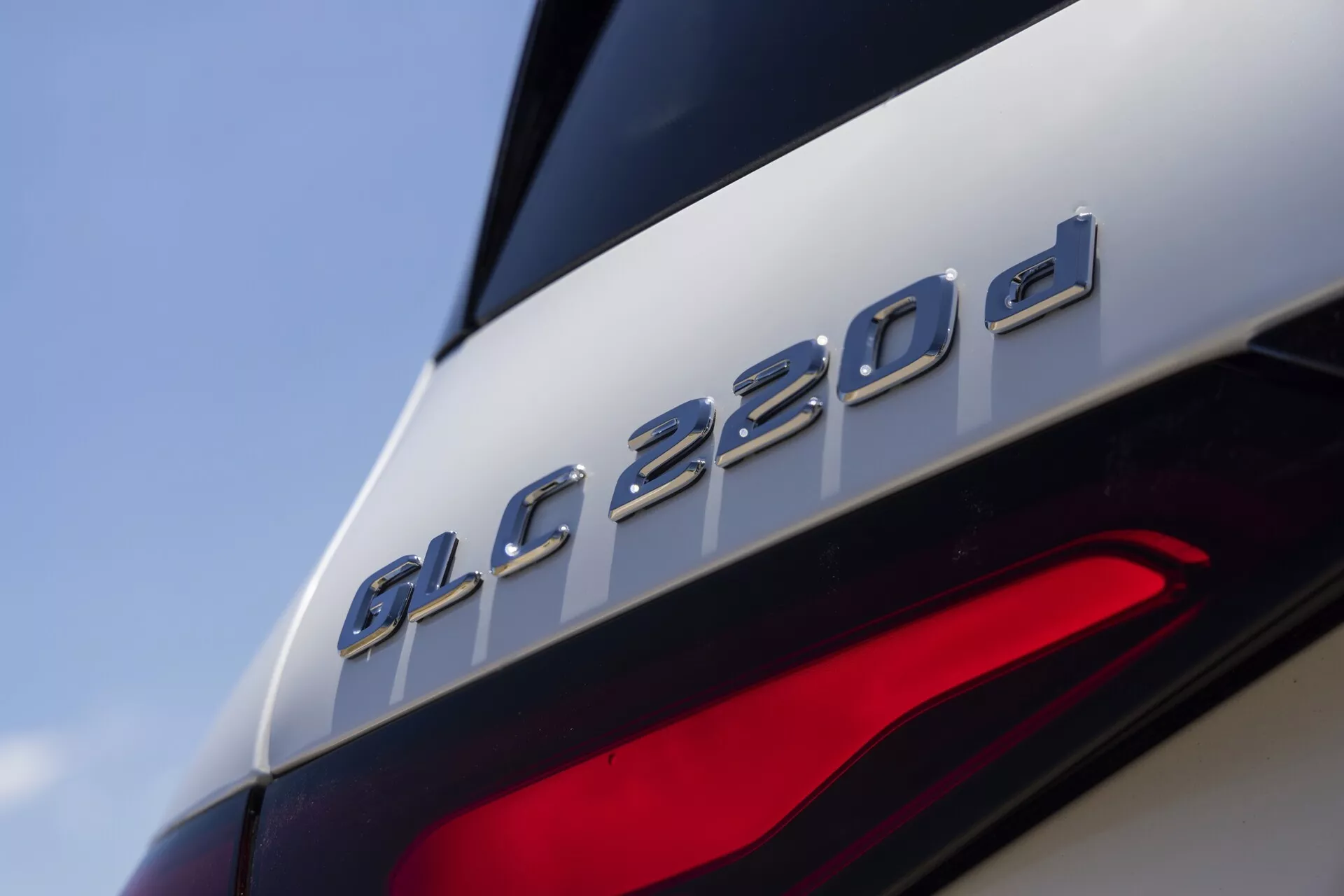 Vue rapprochée de la section arrière de la Mercedes-Benz GLC 220d 2022, mettant en évidence le badge du modèle juste au-dessus du feu arrière partiellement visible. L'image se concentre sur le badge 'GLC 220d', suggérant qu'il s'agit d'une variante diesel. La prise de vue depuis un angle bas, légèrement vers le haut, met en valeur les lignes de design élégantes, la finition métallique de l'insigne contrastant avec la peinture brillante et les éléments LED modernes du feu arrière, indiquant une technologie d'éclairage avancée.
