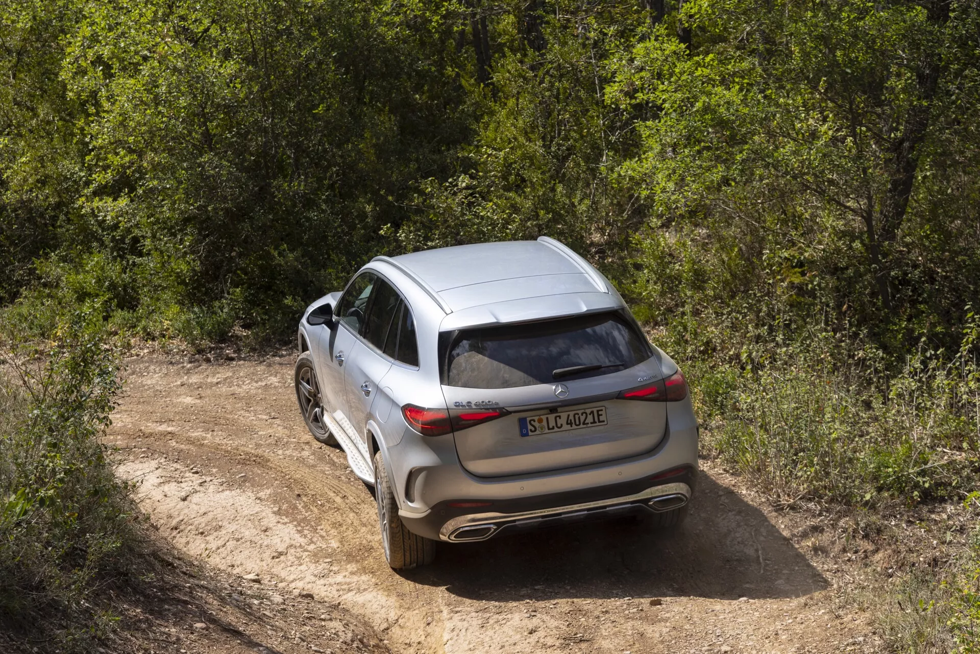 L'image montre un crossover ou SUV moderne et luxueux pris sous un angle surélevé à trois quarts arrière, affichant clairement son arrière et partiellement son profil latéral. Le véhicule présente des feux arrière nets et orientés horizontalement, un hayon fermé, des essuie-glaces arrière visibles et un pare-chocs arrière avec des échappements intégrés. La roue arrière en alliage visible a un design à plusieurs rayons avec des pneus de profil modéré, adaptés au tout-terrain léger. La voiture, de couleur argent ou gris clair métallisé, dispose d'un toit panoramique et de barres de toit. Elle est stationnée sur un chemin de terre étroit entouré de feuillage vert, suggérant un style de vie aventureux en plein air.