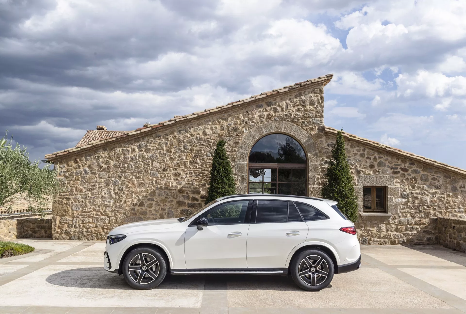 Vue de profil latéral d'un SUV Mercedes GLC 2022 blanc garé sur une surface pavée avec un bâtiment en pierre rustique comprenant une grande fenêtre cintrée en toile de fond. Le ciel couvert offre une lumière naturelle extérieure, avec l'angle de la caméra montrant le toit et les lignes de carrosserie de la voiture du côté passager. Caractéristiques notables : grandes roues en alliage multi-branches, ligne de caractère sous les fenêtres, poignées de portes affleurantes, rails de toit et antenne aileron de requin.