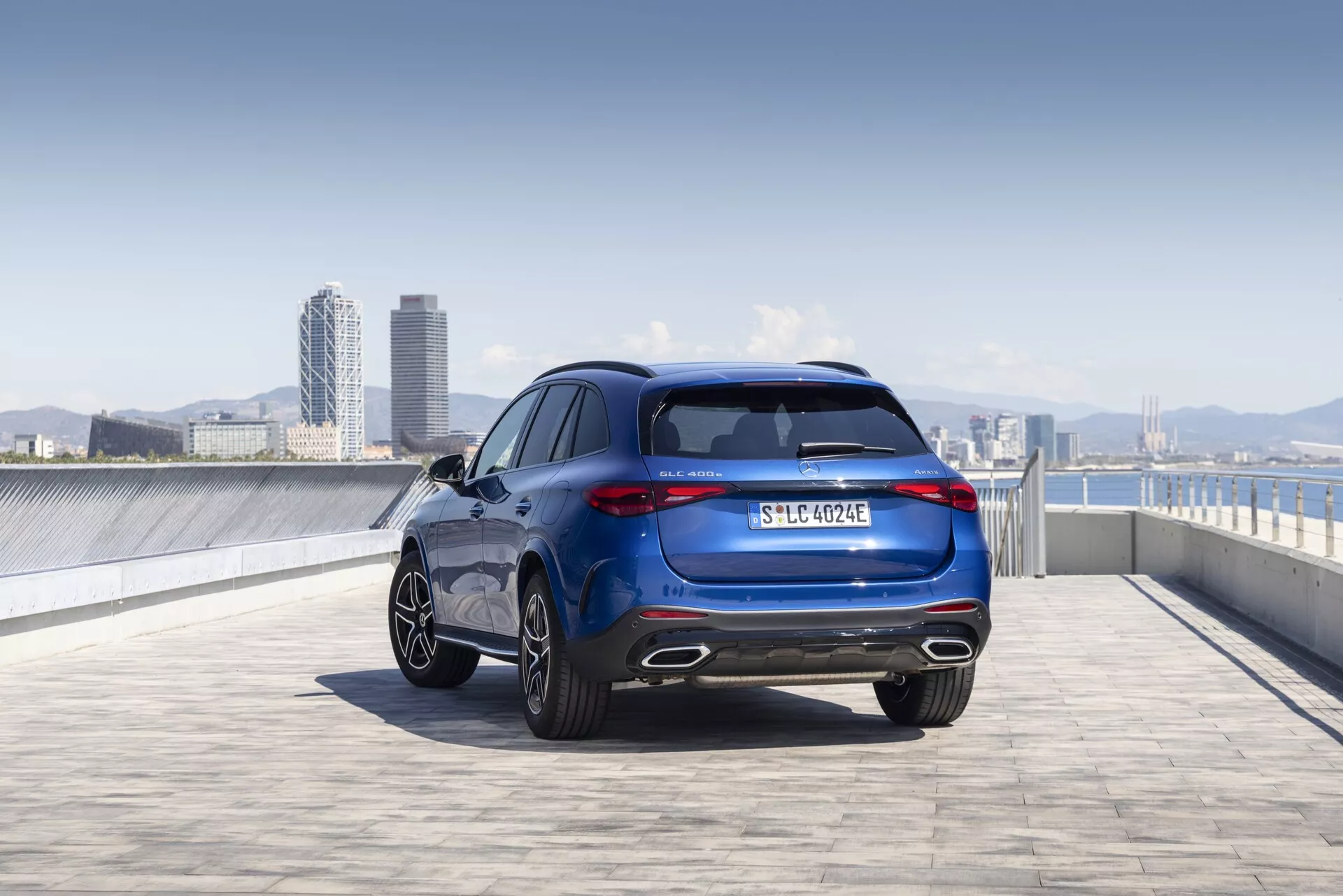 Vue en contre-plongée d'une Mercedes GLC 2022 (254) sous un angle arrière trois quarts, mettant en valeur ses lignes de design élégantes et sportives, ses feux arrière, ses jantes en alliage, sa peinture métallisée et son logo. L'image capture toute la hauteur et la largeur du véhicule, mettant en avant des éléments comme la lunette arrière, le système d'échappement, la plaque d'immatriculation et peut-être un badge de finition ou de transmission. Le cadre extérieur, possiblement un parking sur le toit avec une ligne d'horizon urbaine en arrière-plan, souligne l'attrait moderne et sophistiqué du véhicule.