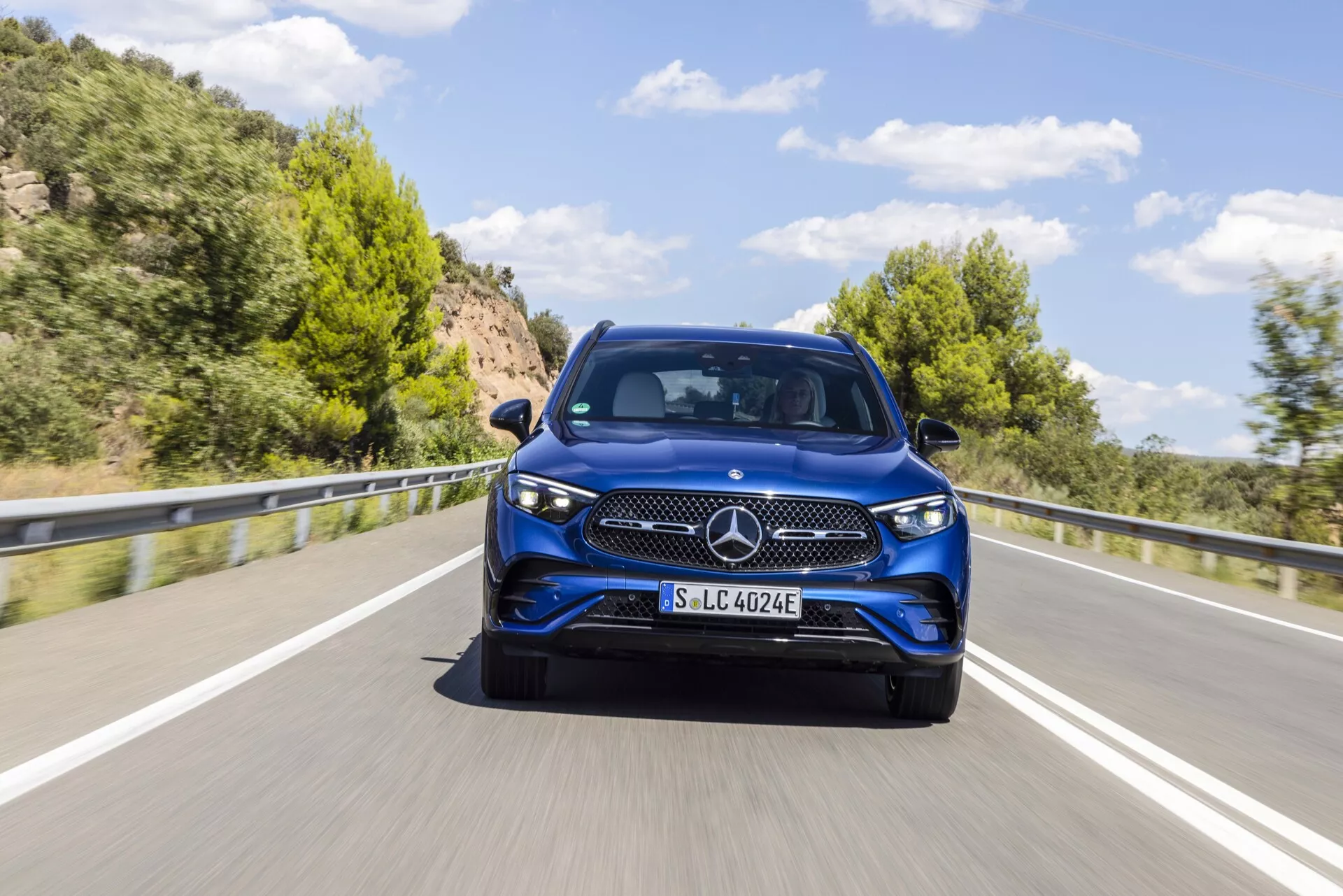 Image d'un modèle bleu Mercedes-Benz GLC 254 de 2022 capturé sous un angle trois quarts avant, mettant en évidence le design élégant, l'emblème étoilé à trois branches, le motif détaillé de la calandre, les phares élégants se fondant dans les ailes et les détails clairs du pare-chocs avant, du capot et des portes. La voiture, photographiée en mouvement sur une route de campagne pittoresque à deux voies avec des collines verdoyantes à l'arrière-plan, dispose probablement d'une technologie électrique ou hybride comme le suggère la marque 'EQ' sur le support de la plaque. L'image est détaillée avec un flou de mouvement indiquant le mouvement et montre les feux de jour de la voiture en journée, sous un ciel clair.
