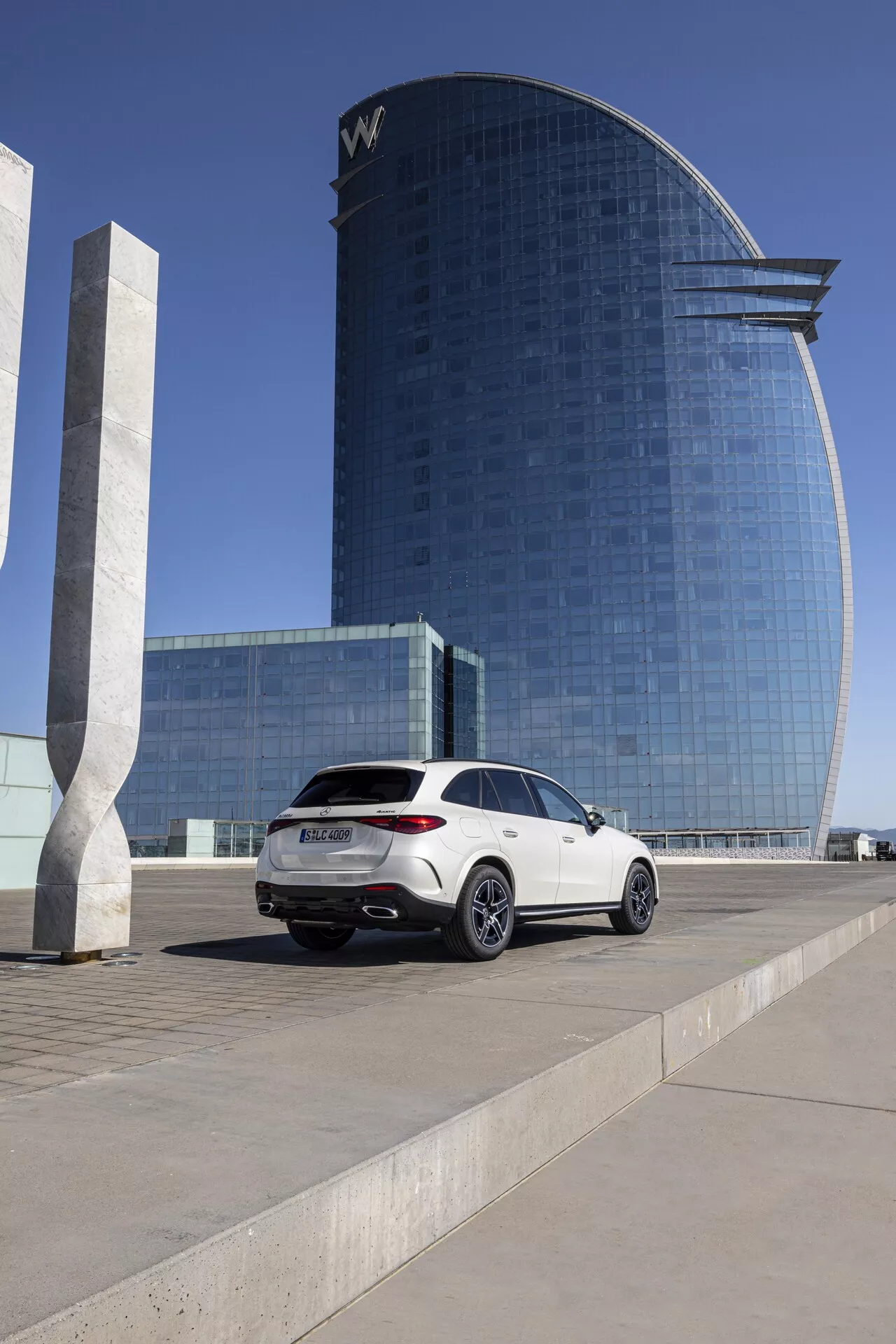 Mercedes GLC 2022 - SUV de luxe blanc stationné sur une place; angle moyen-bas capturant le profil latéral du côté passager et l'arrière, montrant des feux arrière modernes, un double échappement, des roues en alliage multibrins, une ligne de toit de type coupé, des vitres teintées; bâtiment contemporain d'hôtel ou de bureau avec façade en verre réfléchissant en arrière-plan sous un ciel clair et lumineux.