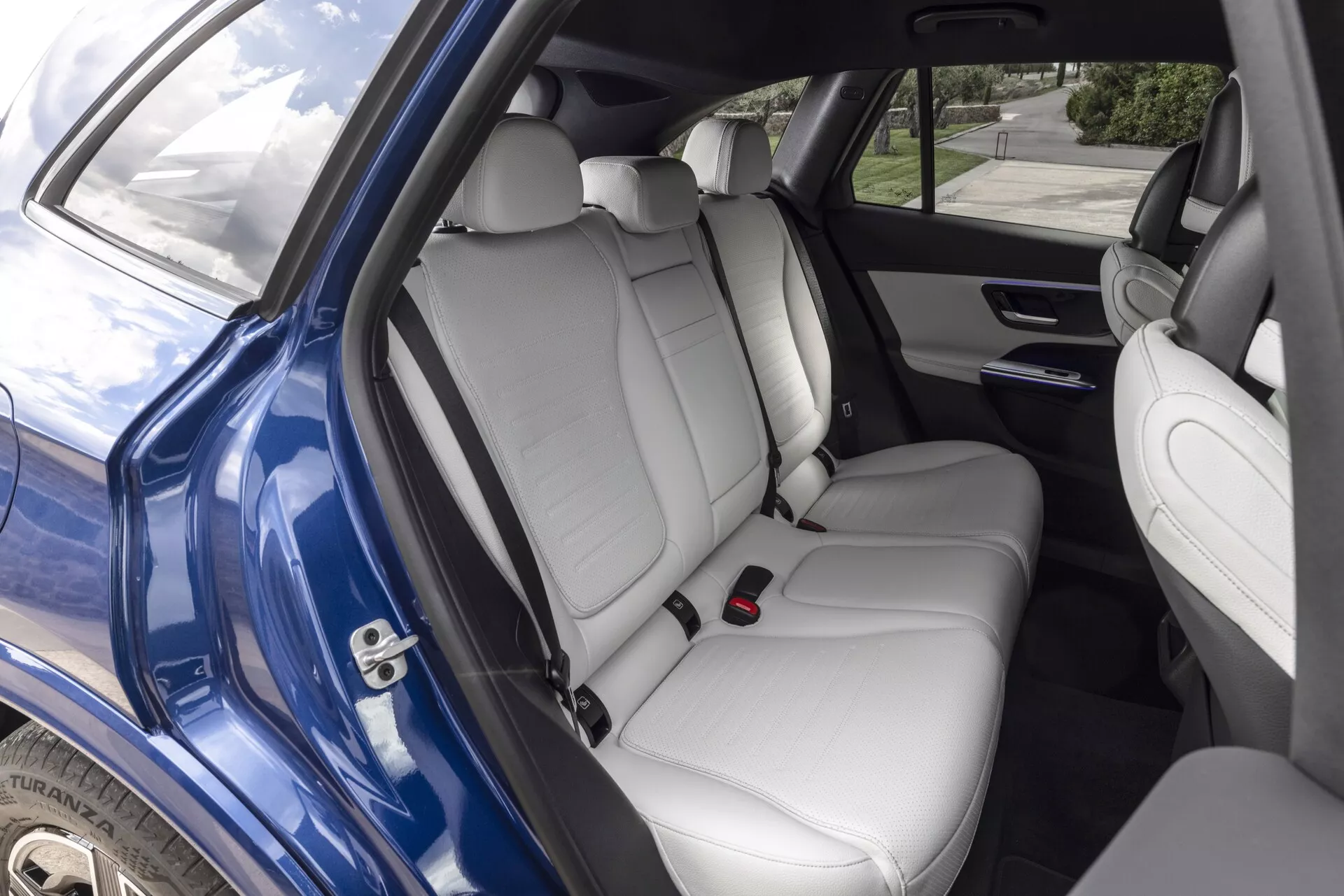 Image de l'intérieur de la Mercedes GLC 2022 montrant le compartiment arrière vu depuis la porte arrière droite ouverte. Les sièges arrière de couleur claire, en cuir, disposent d'appuie-têtes et d'un accoudoir rabattable avec porte-gobelets. La porte arrière contient une unité de haut-parleurs, des commandes de vitres électriques et une poche de rangement. Les dossiers des sièges avant sont dotés de poches de rangement et l'espace généreux de la zone arrière souligne le design familial et les caractéristiques de luxe de la voiture, la lumière naturelle illuminant la scène.