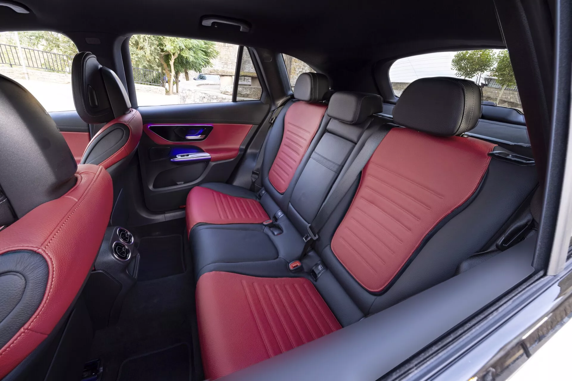 Vue intérieure d'une Mercedes GLC 2022 depuis le côté droit, capturant les sièges arrière avec une luxueuse sellerie bicolore en rouge et noir en cuir ou similicuir, contrastée par des surpiqûres détaillées. Les caractéristiques comprennent un accoudoir central rabattable avec porte-gobelets, des éclairages d'ambiance LED bleus renforçant l'atmosphère moderne de l'habitacle, une sortie d'air pour la climatisation arrière et des haut-parleurs intégrés dans les panneaux de porte, démontrant un éclairage avancé, des finitions de luxe et le confort des passagers.
