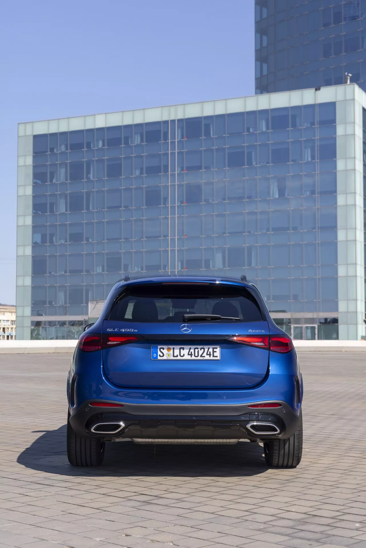 Vue arrière de la Mercedes GLC 2022 (254) en bleu métallique profond, positionnée en extérieur dans un environnement urbain avec un immeuble à façade vitrée réfléchissante en arrière-plan. La voiture est centrée, capturée sous un angle légèrement bas, mettant en valeur le coffre, le pare-chocs arrière, les feux arrière LED, les jantes en alliage et le spoiler de toit. Les caractéristiques incluent un badge central proéminent, la plaque d'immatriculation, la désignation du modèle à gauche, 'E' indiquant un modèle électrique ou hybride à droite, et un diffuseur arrière avec sorties d'échappement, suggérant un modèle hybride.