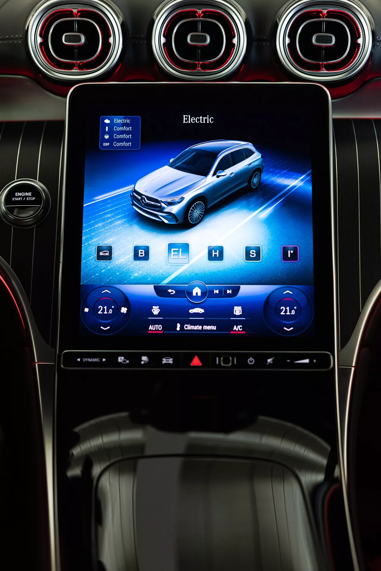 Vue intérieure du tableau de bord d'une voiture avec le système d'infodivertissement Mercedes GLC 2022. Le point focal est un écran tactile orienté verticalement intégré dans le tableau de bord. L'interface vibrante montre une représentation graphique d'un véhicule électrique bleu en légère perspective 3/4 avant. Le mot 'Electric' est affiché en haut de l'écran, indiquant le type de motorisation du véhicule. L'écran inclut également des réglages de contrôle climatique à deux zones avec une température réglée sur '21' de chaque côté, ainsi que des boutons de réglage plus et moins, et diverses icônes en bas. Les matériaux du tableau de bord entourant l'écran semblent inclure des plastiques brillants et texturés et peut-être des accents en cuir. Au-dessus de l'écran se trouvent des bouches d'aération circulaires avec des accents rouges, ajoutant une esthétique sportive ou haut de gamme à l'intérieur de la voiture.