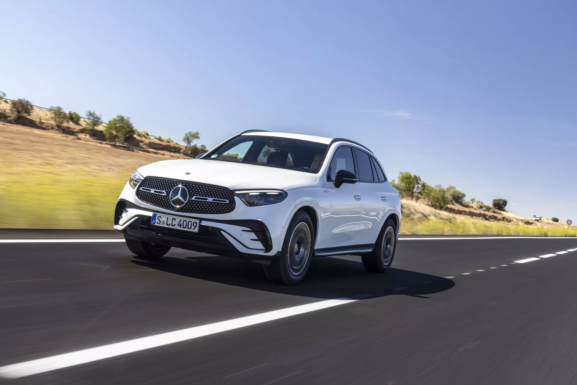 Prise de vue dynamique du SUV de luxe Mercedes GLC 2022 avançant sur une route en asphalte lisse, montrant son profil avant et latéral. Les fonctionnalités incluent la grille emblématique à étoile à trois branches, les élégants phares à LED, les capteurs visibles sur le pare-chocs avant indiquant des systèmes avancés d'assistance à la conduite, les lignes sculptées du capot, les jantes en alliage, les rétroviseurs latéraux aérodynamiques avec clignotants intégrés et une ligne de toit fluide pour une silhouette plus sportive. La posture dominante du véhicule et le léger flou de mouvement autour des roues soulignent son mouvement. Fond de ciel clair et paysage désertique.