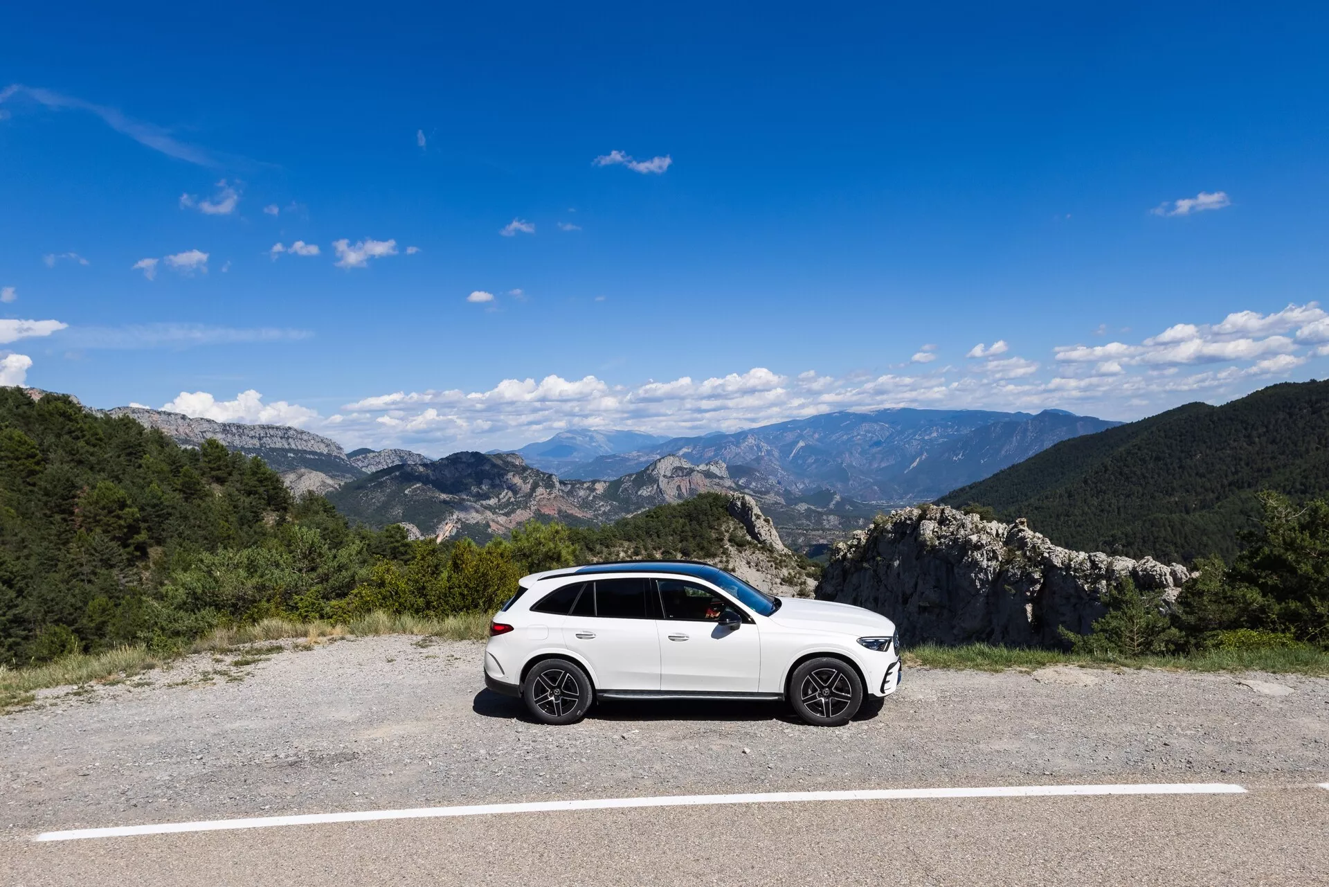 Un SUV sportif Mercedes GLC 2022 blanc est garé sur une zone pavée plate avec un paysage montagneux naturel en arrière-plan, sous un ciel bleu clair. La vue de profil latéral montre le côté conducteur du véhicule, mettant en valeur des éléments de design tels que les roues en alliage, les rétroviseurs latéraux, les garnitures des fenêtres et les lignes de carrosserie. L'éclairage et la clarté permettent de voir les contours subtils des portes latérales.