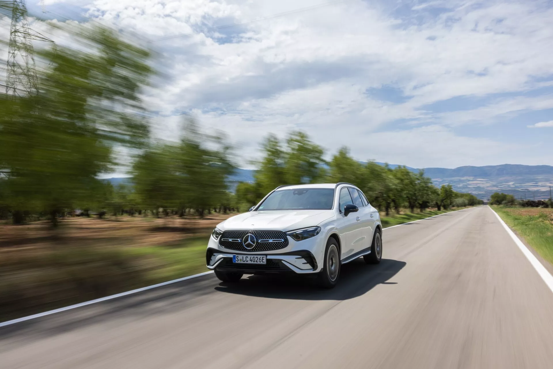 Vue avant trois quarts d'une Mercedes GLC 2022 blanche en mouvement sur une route à deux voies, mettant en avant sa calandre avant, ses phares, une partie du capot, le pare-brise et le côté conducteur. L'arrière-plan est flou en raison du mouvement, soulignant la vitesse de la voiture. Le design élégant, la calandre fermée, les phares LED économes en énergie et les feux de jour visibles, ainsi qu'un paysage moderne avec des arbres flous et un terrain vallonné, présentent un aspect dynamique et stylé idéal pour les supports promotionnels.
