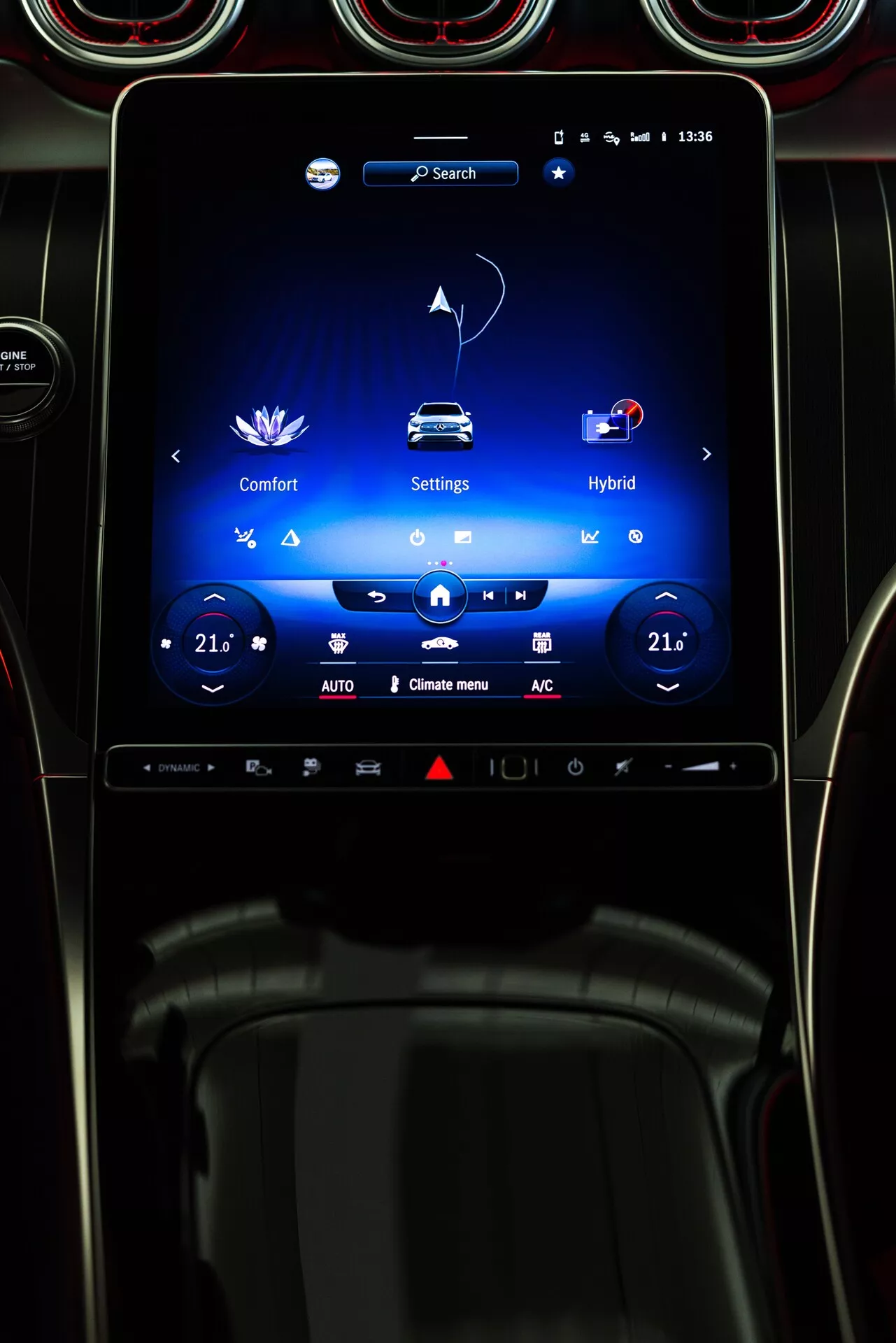 L'image montre l'intérieur d'une Mercedes GLC 2022, en mettant l'accent sur l'écran tactile d'infodivertissement central orienté verticalement sur le tableau de bord. L'écran affiche les menus 'Comfort,' 'Settings' et 'Hybrid,' indiquant des fonctionnalités de confort réglables et un groupe motopropulseur hybride. Les commandes de climatisation bizone sont réglées à 21 degrés Celsius. La zone environnante comprend des évents circulaires avec une finition métallique, ajoutant une touche luxueuse. La photo est prise de la perspective du conducteur, mettant en avant des fonctionnalités technologiques et de connectivité avancées.