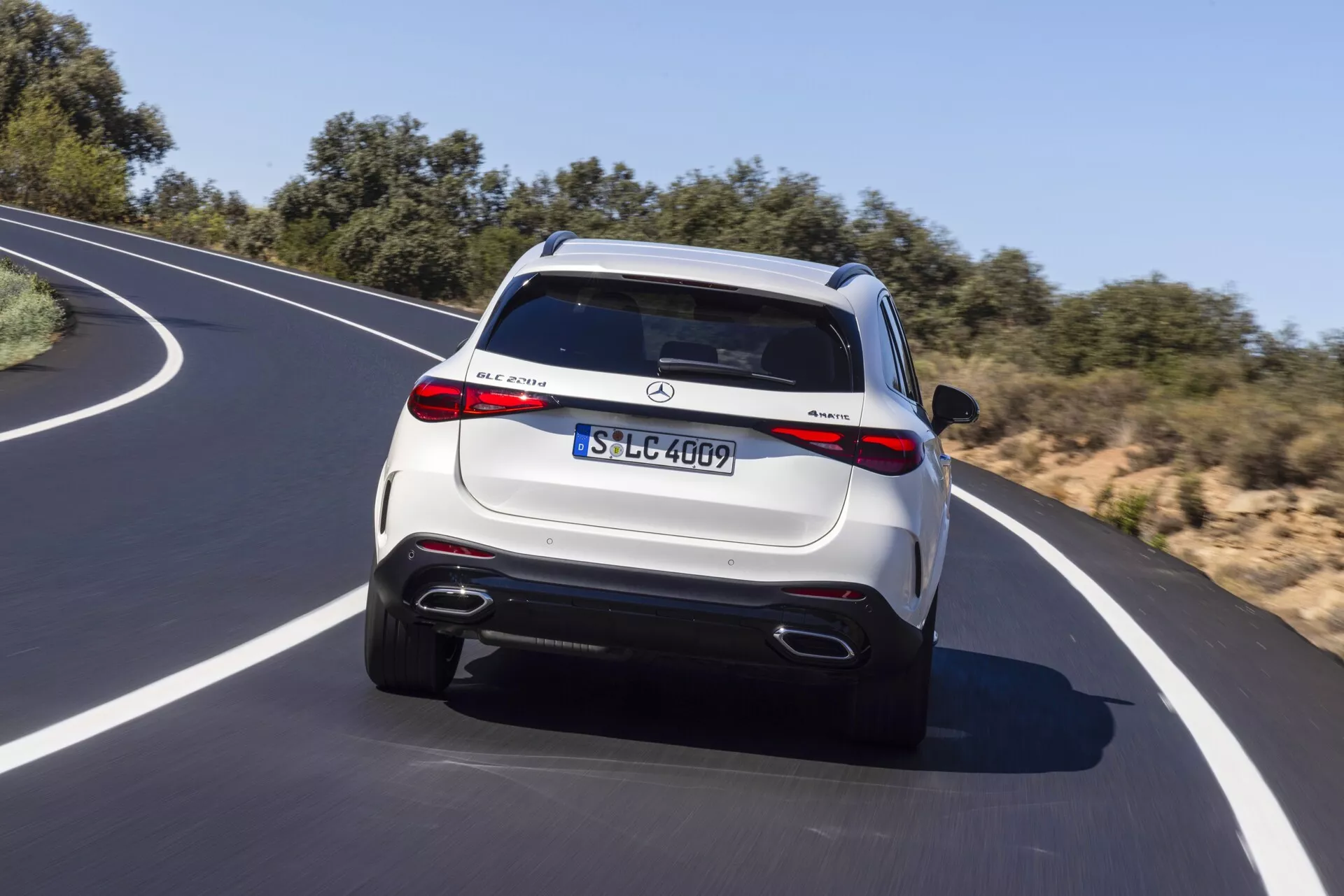 Vue arrière d'un SUV Mercedes-Benz GLC 2022 en mouvement sur une route asphaltée sinueuse sous un ciel clair. Le véhicule présente une finition métallique claire, des feux arrière à LED, des doubles sorties d'échappement, un pare-chocs arrière de type diffuseur et un spoiler de toit avec feu stop intégré. Des badges bien visibles et une plaque d'immatriculation de style européen sont également visibles, indiquant les détails du modèle et du niveau de finition. Des jantes à multiples rayons et des caractéristiques technologiques potentielles comme des capteurs de stationnement et une caméra de recul ajoutent à son attrait luxueux.