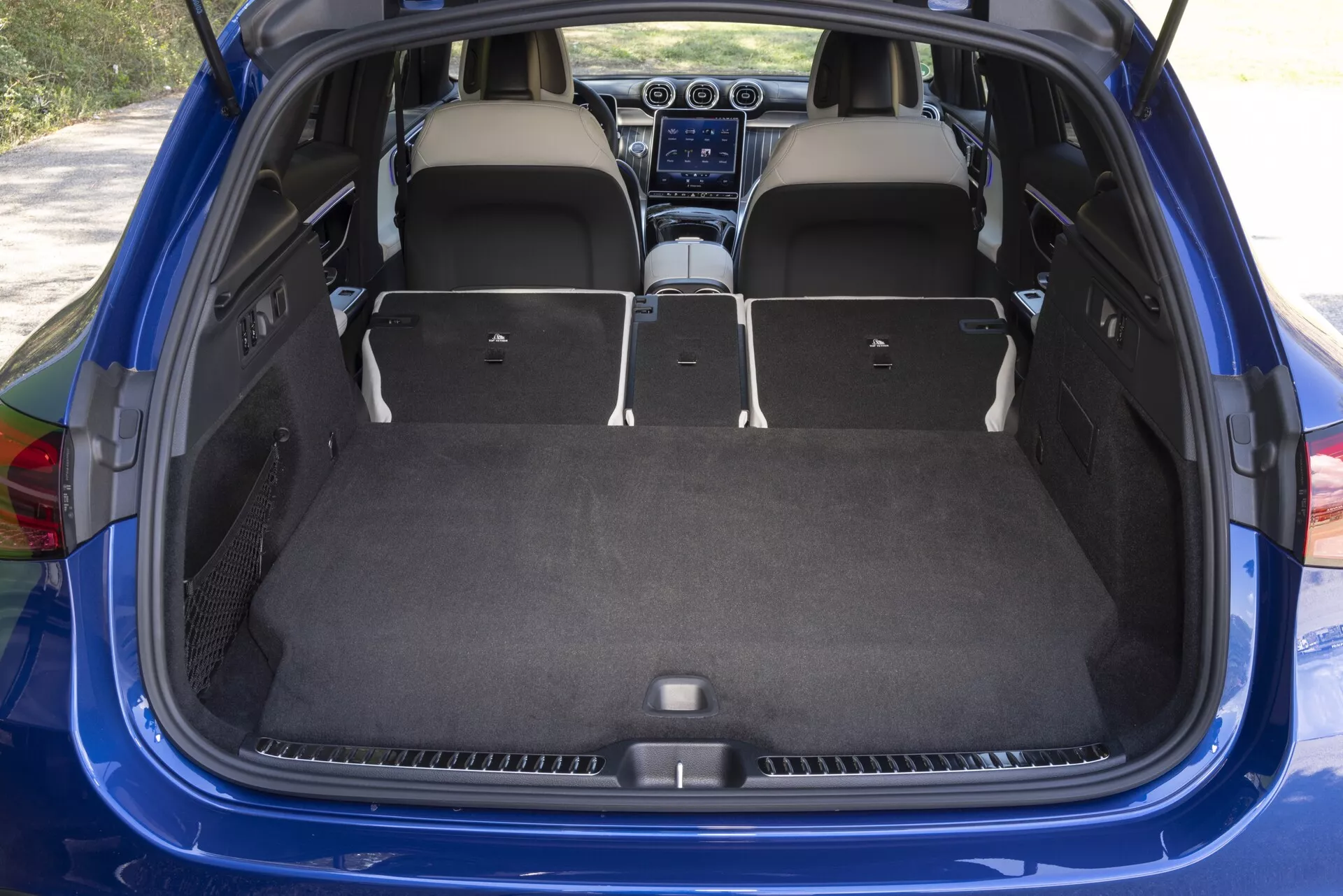 Vue intérieure de l'espace de chargement du Mercedes GLC 2022 (254) avec le hayon ouvert, montrant un compartiment de charge spacieux et son design. Les sièges arrière sont rabattus, révélant une caractéristique de siège arrière fractionnable 60/40 pour agrandir l'espace de chargement. Plancher de chargement plat pour un chargement et déchargement faciles, moquette en bon état suggérant un intérieur neuf ou bien entretenu. Éclairage par lumière naturelle indiquant une photo prise pendant la journée. Aperçu de l'habitacle avec console centrale, système d'infodivertissement et bouches de climatisation indiquant un écran tactile et la climatisation. Tissu d'ameublement clair et intérieurs de portes alignés avec l'esthétique globale du véhicule. Couleur bleue visible sur les bords de l'ouverture du hayon.