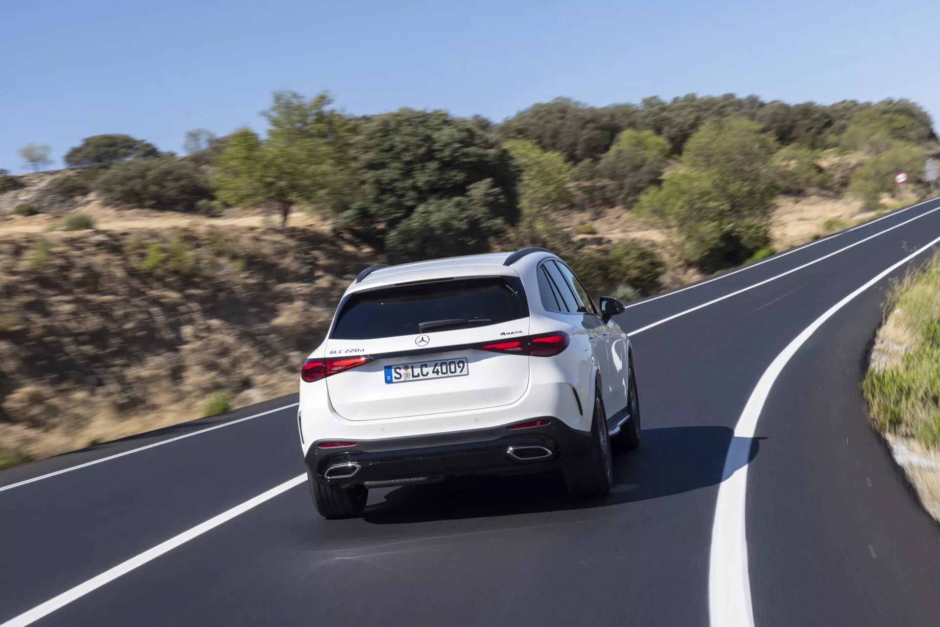 Vue arrière d'un SUV Mercedes GLC 2022 moderne en mouvement, présentant des doubles sorties d'échappement, un diffuseur arrière aérodynamique, des feux arrière à LED, un aileron arrière sportif et des caractéristiques pratiques telles qu'un essuie-glace arrière et un troisième feu stop élevé. La peinture de couleur claire du SUV, comme le blanc ou l'argent, rehausse son apparence haut de gamme tout en roulant sur une route asphaltée bordée de végétation naturelle. Le flou de mouvement met en évidence les performances dynamiques du véhicule.