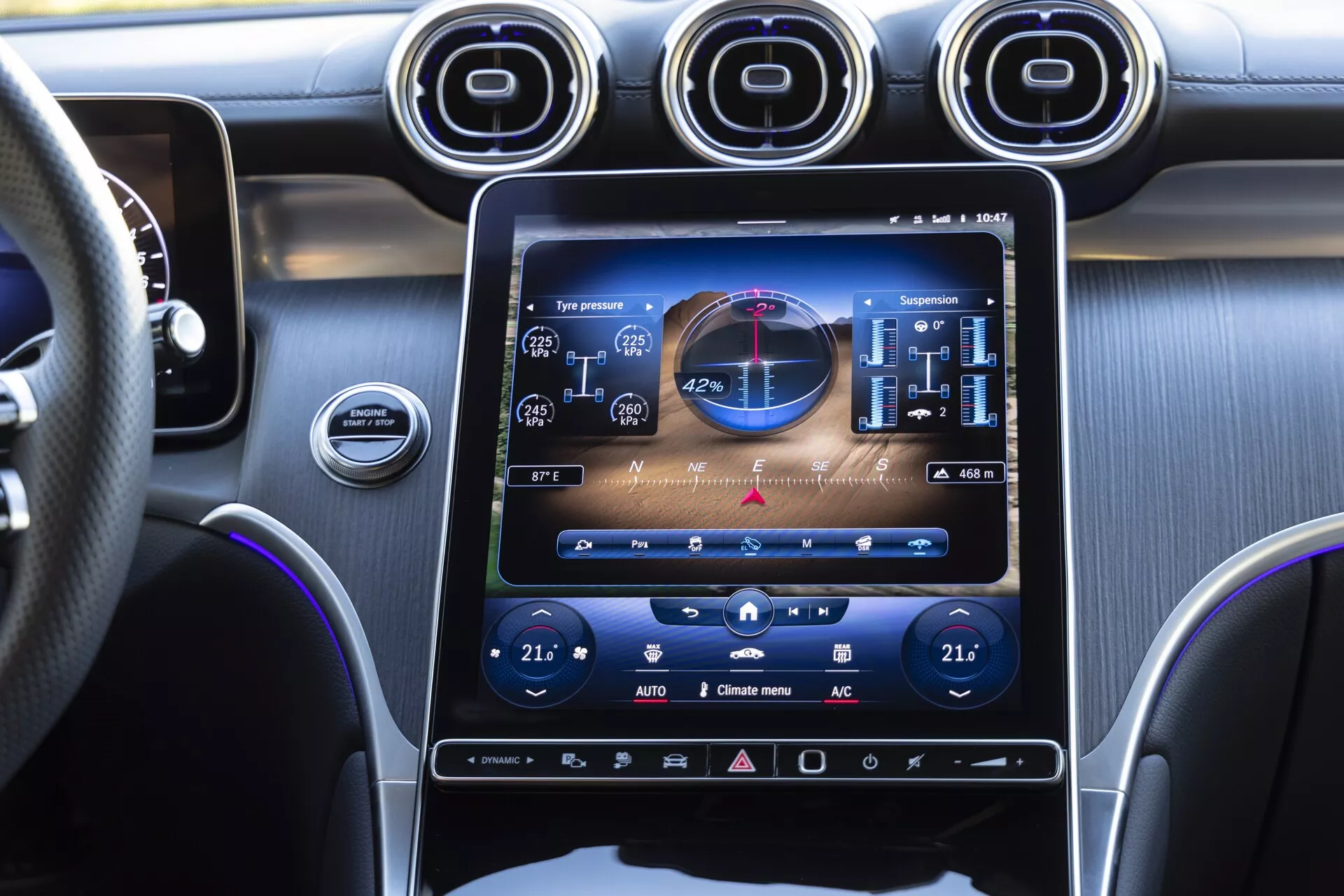 Vue intérieure en gros plan du tableau de bord d'une Mercedes GLC 2022 depuis le côté passager, mettant en avant le grand écran tactile d'infodivertissement en orientation portrait affichant les fonctions du véhicule, les réglages de la climatisation bi-zone et des suspensions personnalisées. Comprend trois bouches d'aération circulaires métalliques au-dessus de l'écran, un volant en cuir avec des surpiqûres, des tons sombres avec des finitions métalliques brossées et des commandes haptiques supplémentaires sous l'écran, soulignant un design moderne et luxueux.