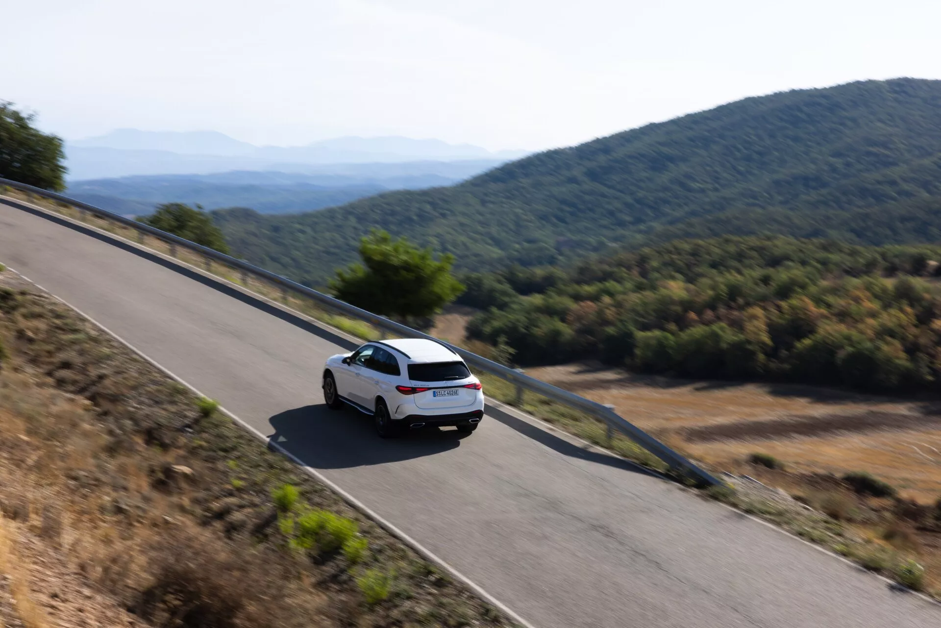 Vue arrière en trois quarts d'un angle élevé d'une Mercedes GLC 2022 (254) blanche moderne conduisant sur une route montagneuse. La voiture, présentant un pare-brise arrière clair, des feux arrière et une partie du coffre, capture une sensation de mouvement avec un léger flou, entourée d'un éclairage naturel et chaud soit le matin soit en fin d'après-midi.